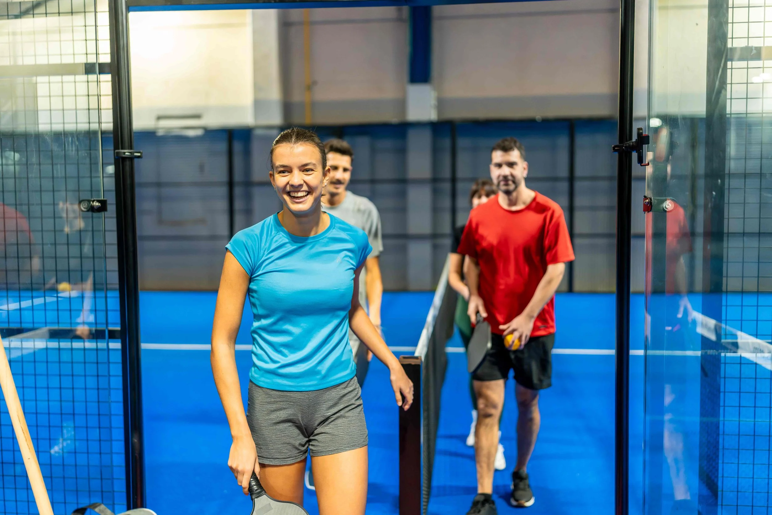 A group of people in athletic clothing standing inside an indoor sports facility, with a woman in a blue shirt smiling and holding a paddle, and others in the background holding pickleball paddles.