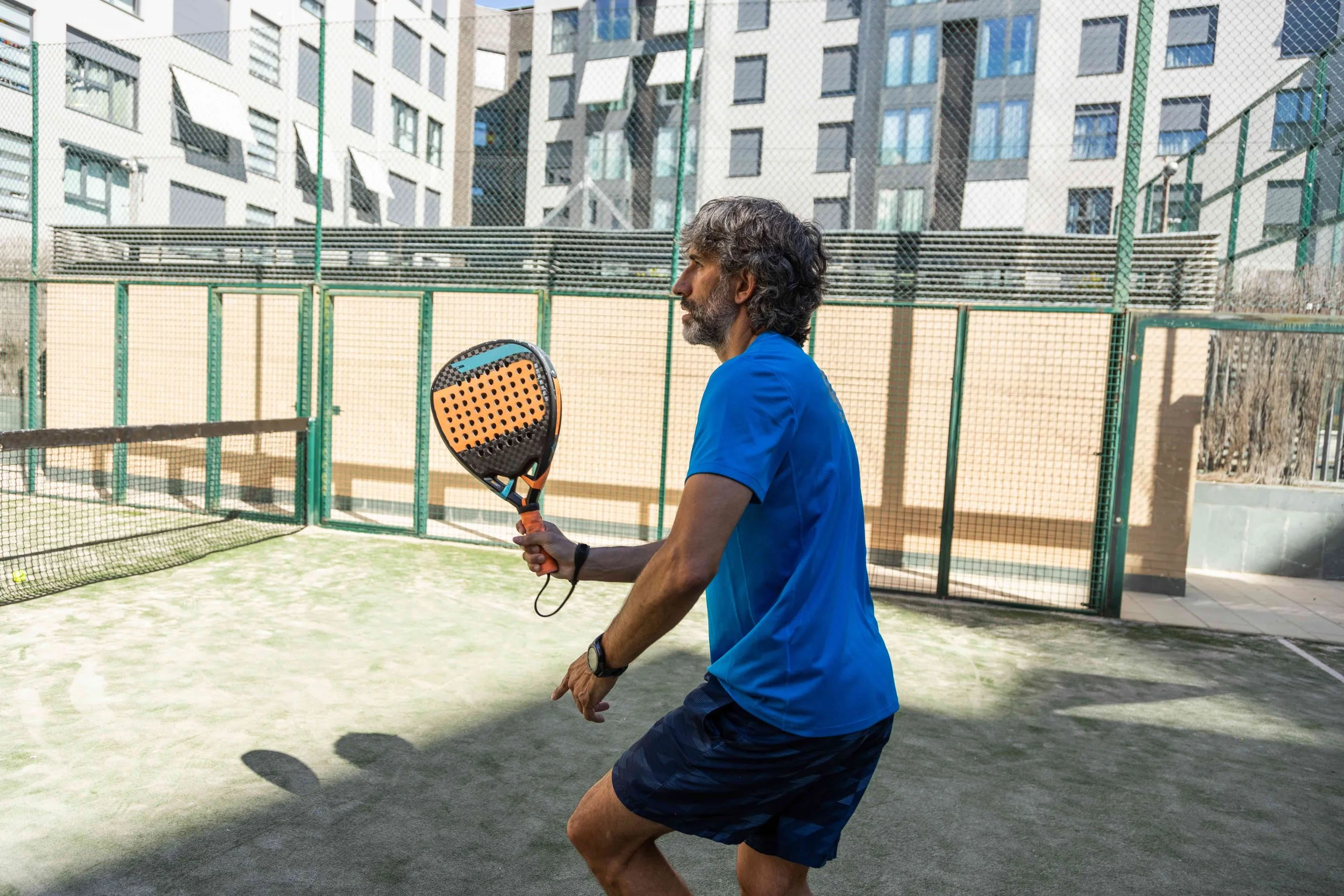 A man with gray hair and a beard wearing a blue shirt and black shorts playing paddle tennis on an outdoor court with a net and urban buildings in the background.