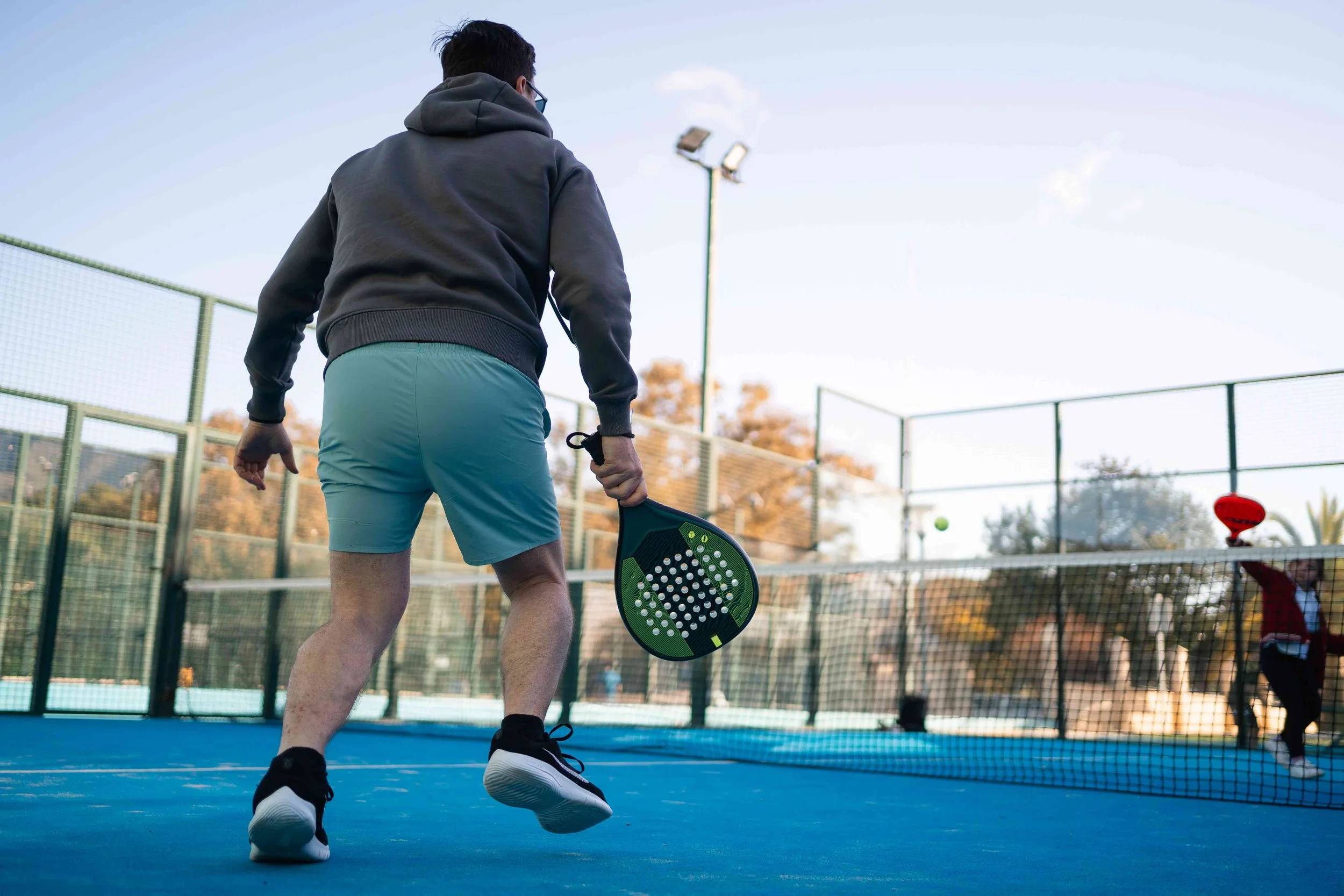 A man playing pickleball on an outdoor court, holding a paddle, with a ball in mid-air, surrounded by a fence and trees.