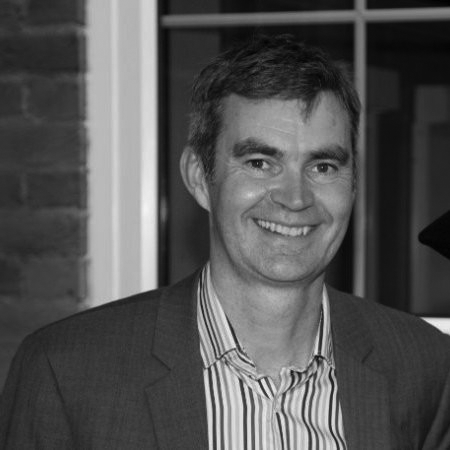 A man with short hair wearing a suit and striped shirt, smiling, standing indoors next to a window and brick wall.