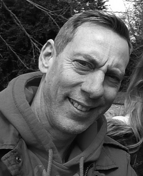 A smiling man with short hair in an outdoor setting, wearing a jacket.