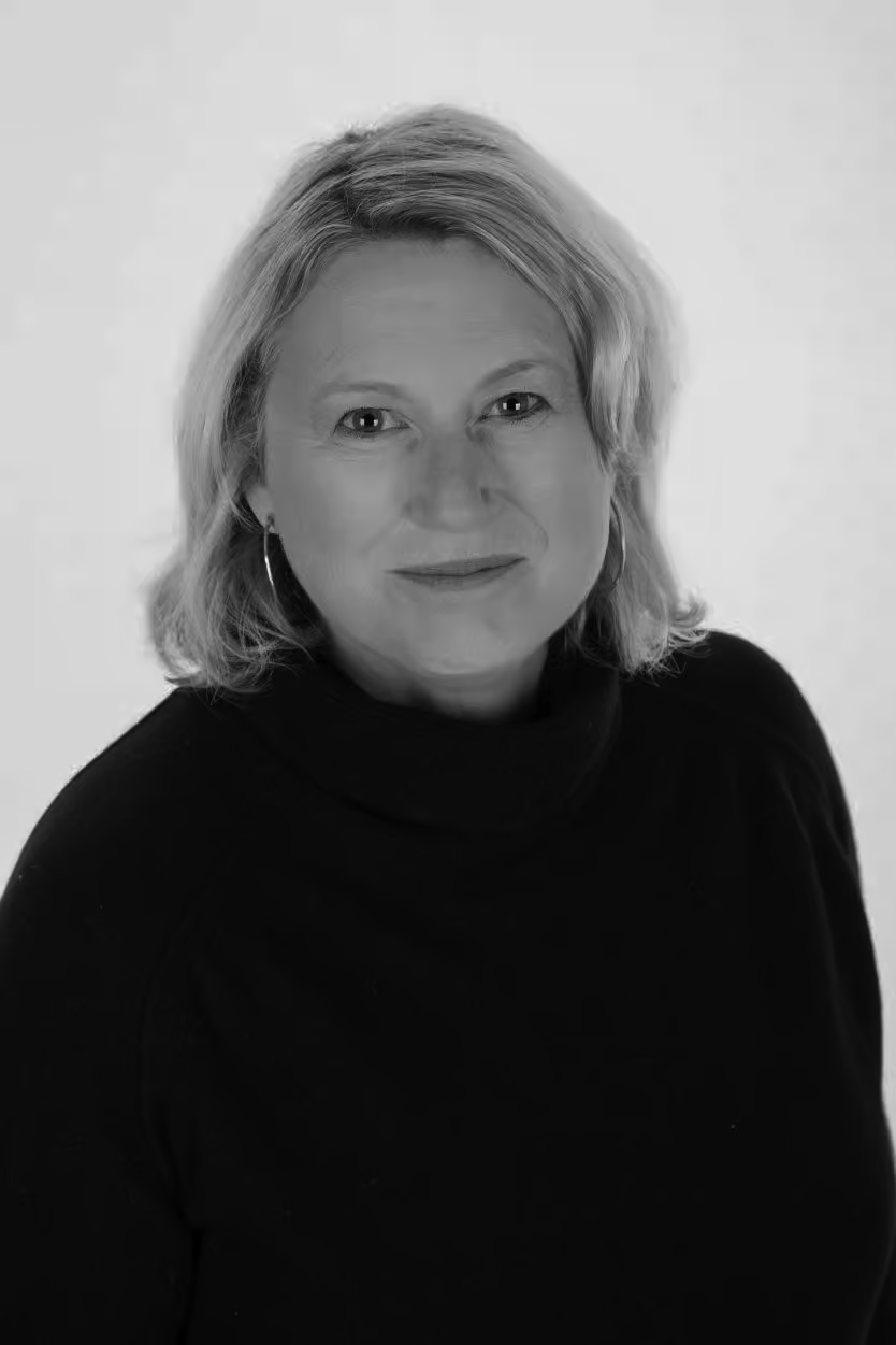 A black and white portrait of a middle-aged woman with shoulder-length light-colored hair, wearing hoop earrings and a dark top, looking at the camera with a slight smile.