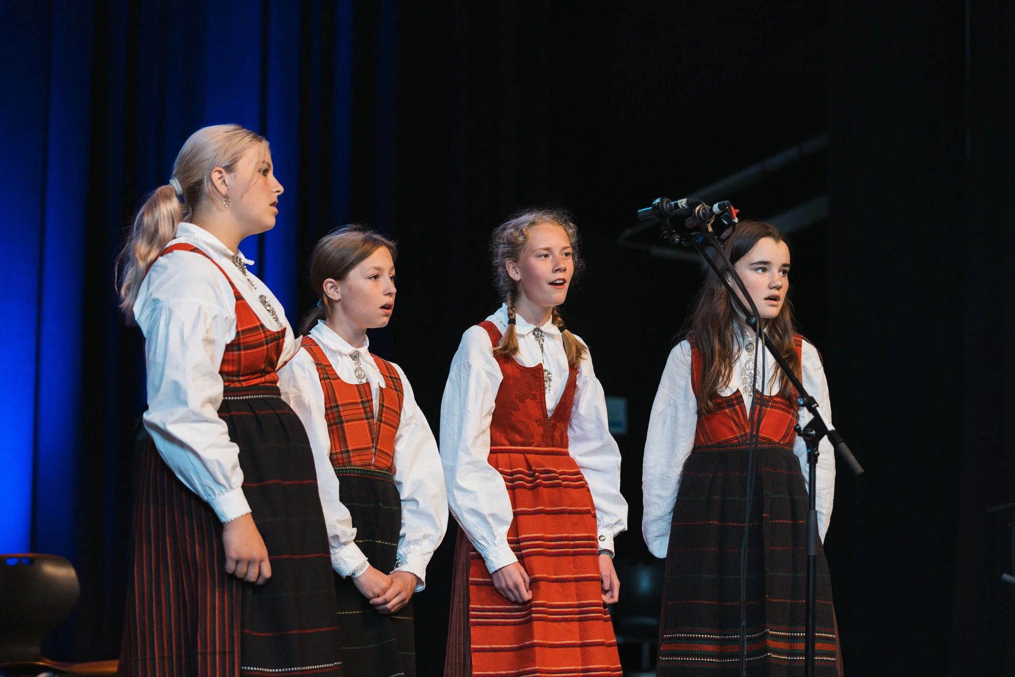 Fire unge jenter synger på en scene iført tradisjonelle norske bunader.