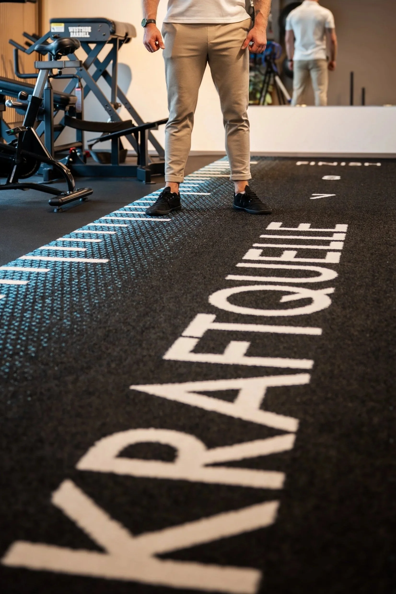 Person steht auf einem schwarzen Laufband im Fitnessstudio mit dem Schriftzug "YAYELOPE TTE" auf dem Boden, im Hintergrund Geräte und eine weitere Person.