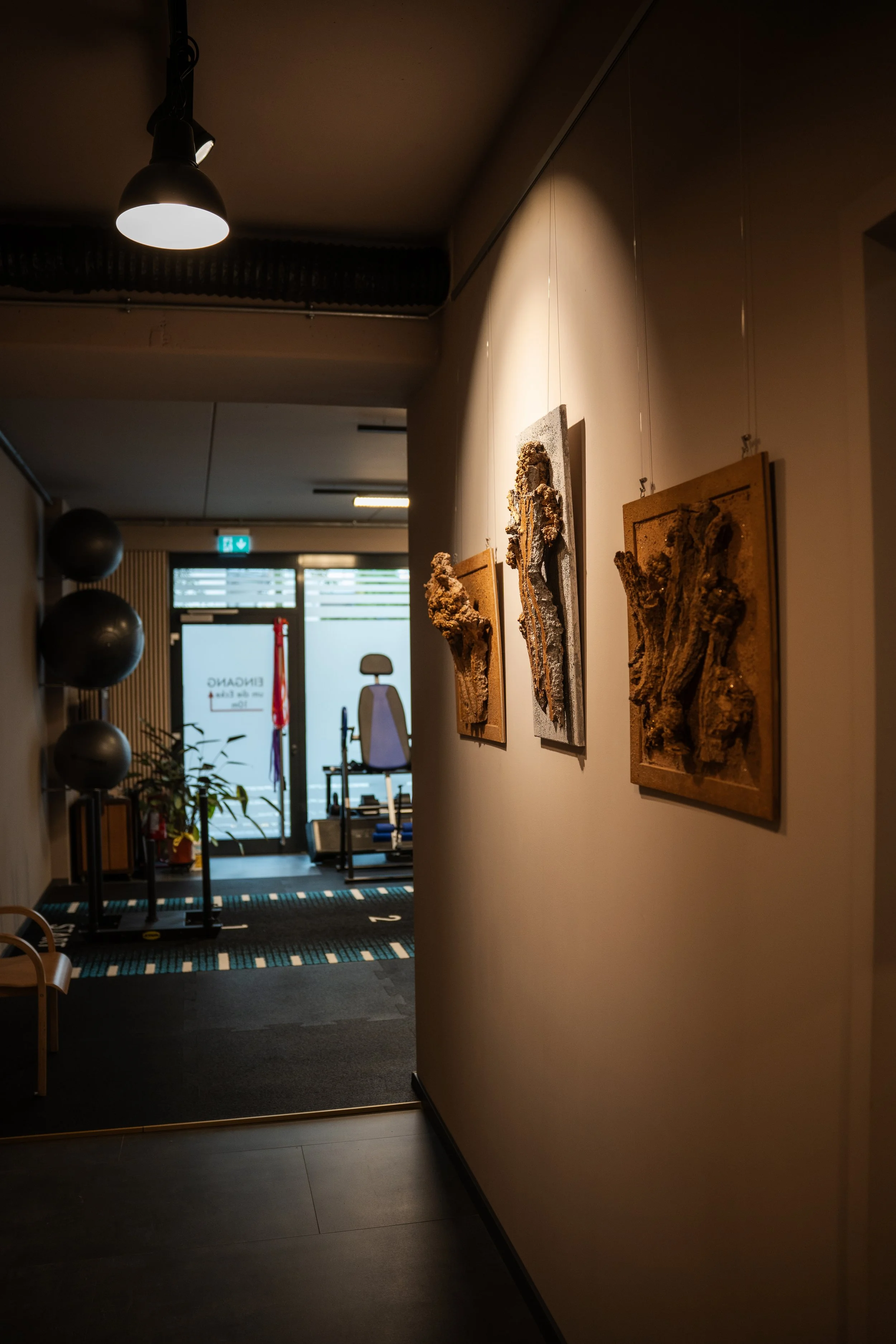Bilder einer Galerie mit drei abstrakten Kunstwerken an einer Wand, beleuchtet durch eine Deckenleuchte, im Hintergrund ein Raum mit Fitnessgeräten und einem Natursteinboden.