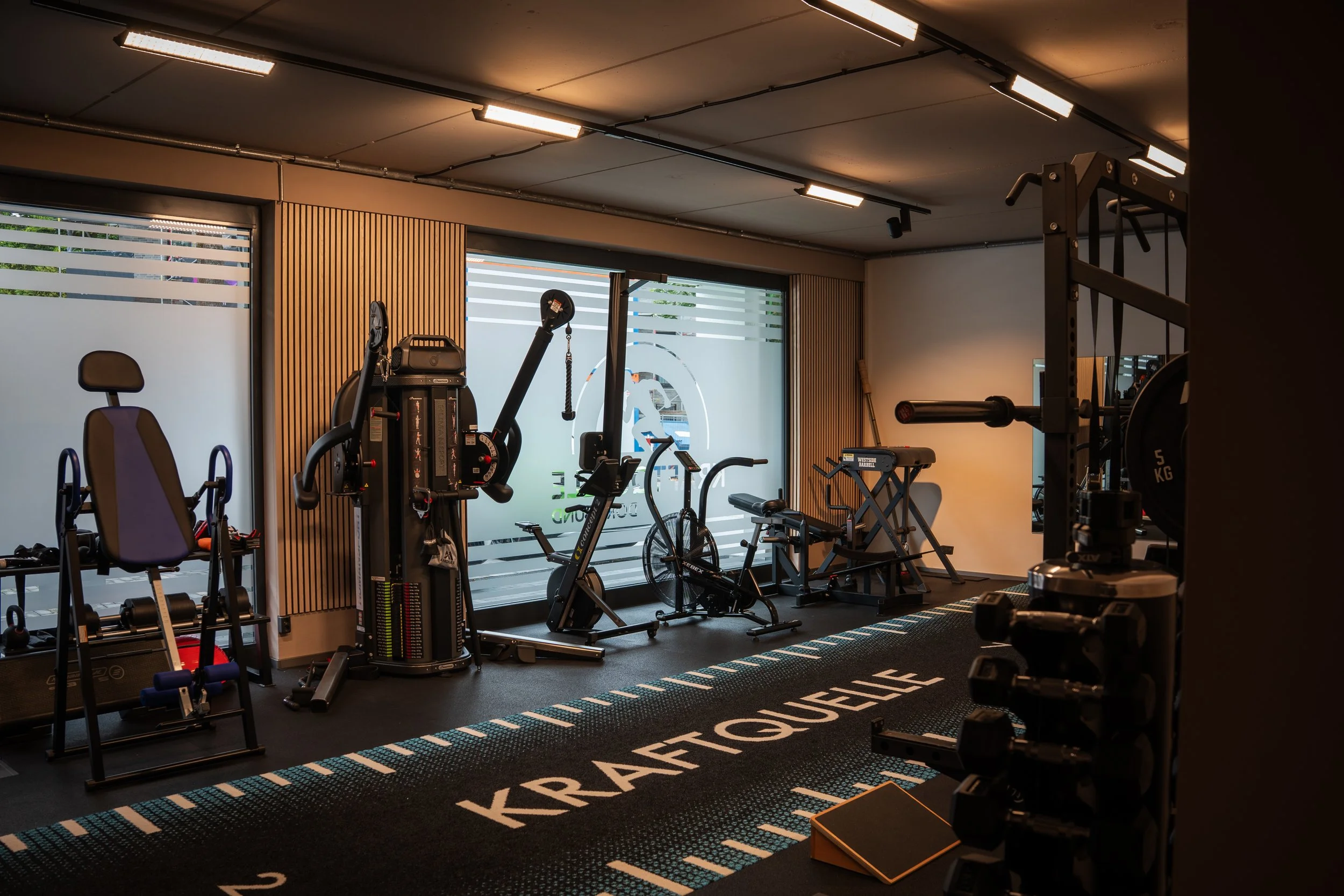 Ein Fitnessraum mit verschiedenen Trainingsgeräten, einschließlich Indoor-Bike, Hanteln, Kraftmaschine und Hantelbänke, auf einem schwarzen Boden mit weißen und blauen Linien und dem Wort ‚KRAFTQUELLE‘.