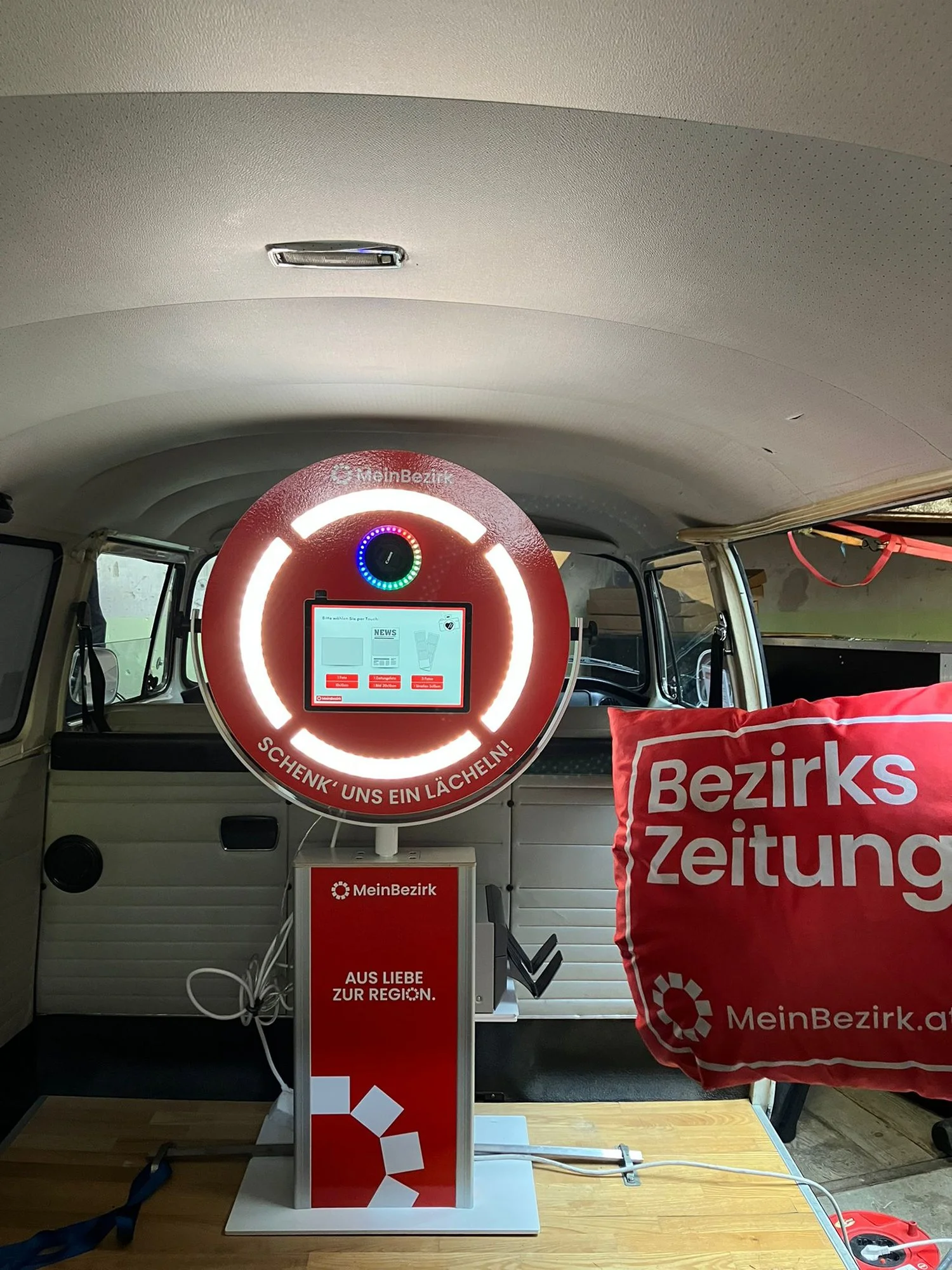 Ein rotes Lichtrunddisplay auf einem kleinen Stand mit dem Text »Schenk' uns ein Lächeln!« und »MeinBezirk«, daneben eine rote Tasche mit weißen Schriftzug »Bezirks Zeitung«.