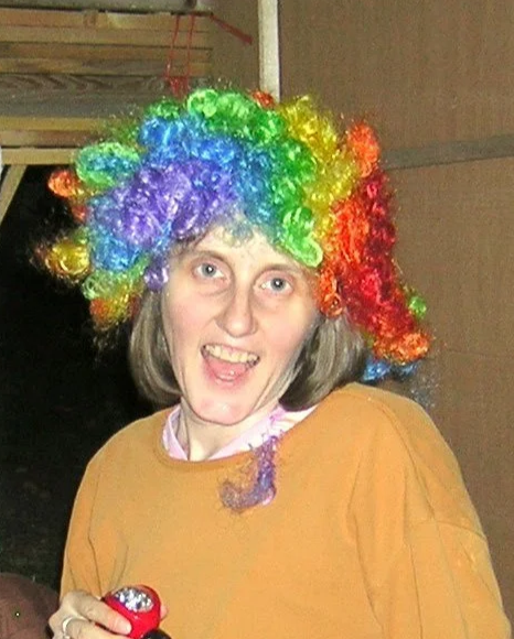 Charlene Clough  wearing a colorful rainbow clown wig, smiling and holding a small red object.