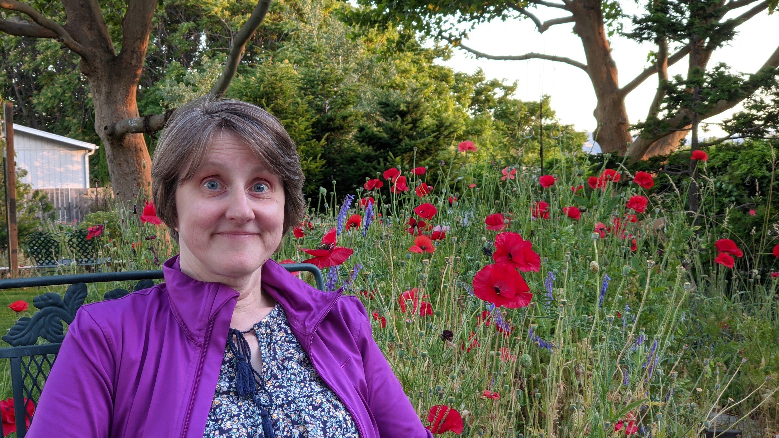Charlene Collings - Clough with short brown hair and blue eyes, wearing a purple jacket and a patterned shirt, sitting on a park bench in a garden lush with colorful red, purple, and green plants, with large trees in the background.