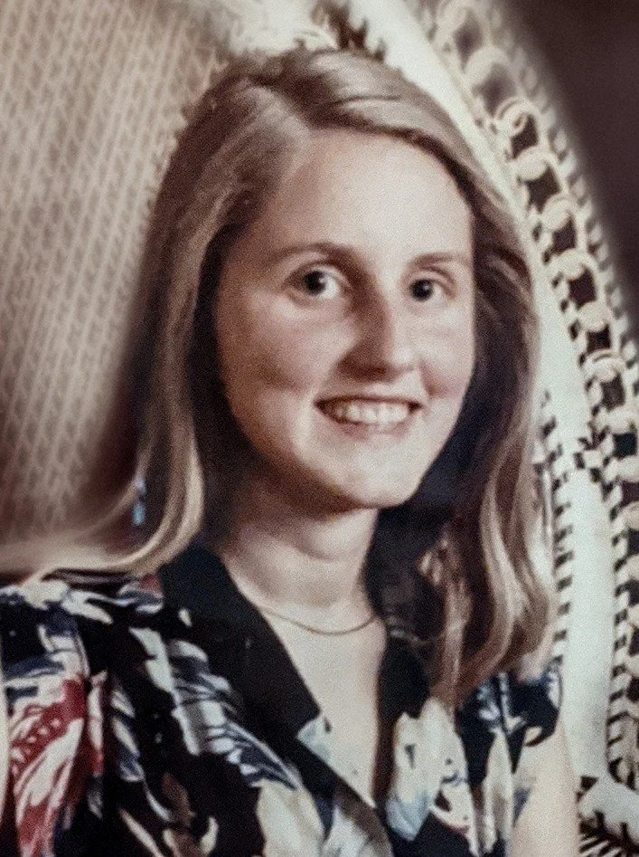 Charlene Clough with shoulder-length blonde hair smiling, wearing a patterned blouse and a delicate necklace, sitting in front of a decorative, carved wooden background.