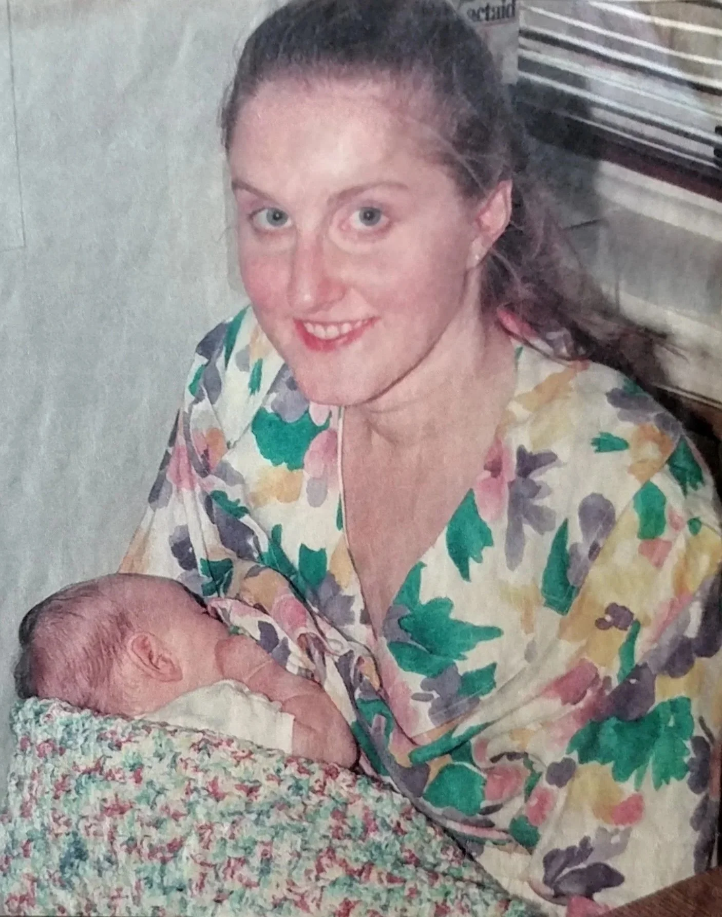 Charlene Clough  in a floral blouse smiling while holding her new daughter, who is lying on a colorful blanket.