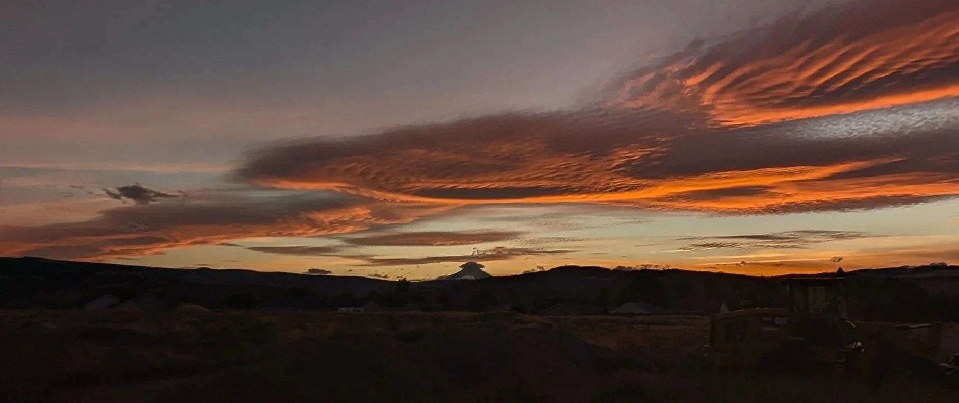 Sunset sky with orange and dark clouds over a hilly landscape with a tractor in the foreground.