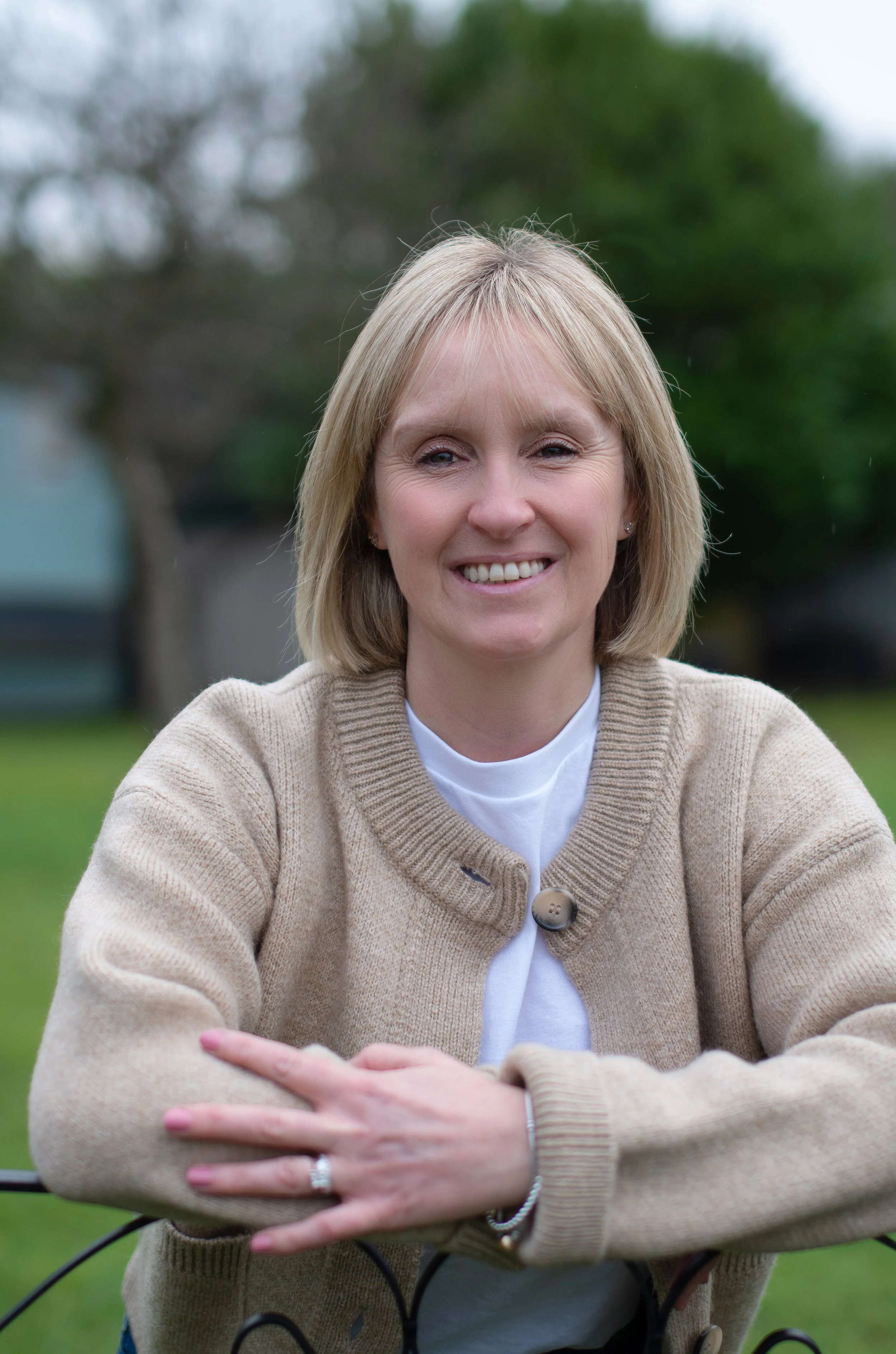 A smiling woman with short blonde hair, wearing a beige cardigan over a white shirt, sitting outdoors in front of green trees.