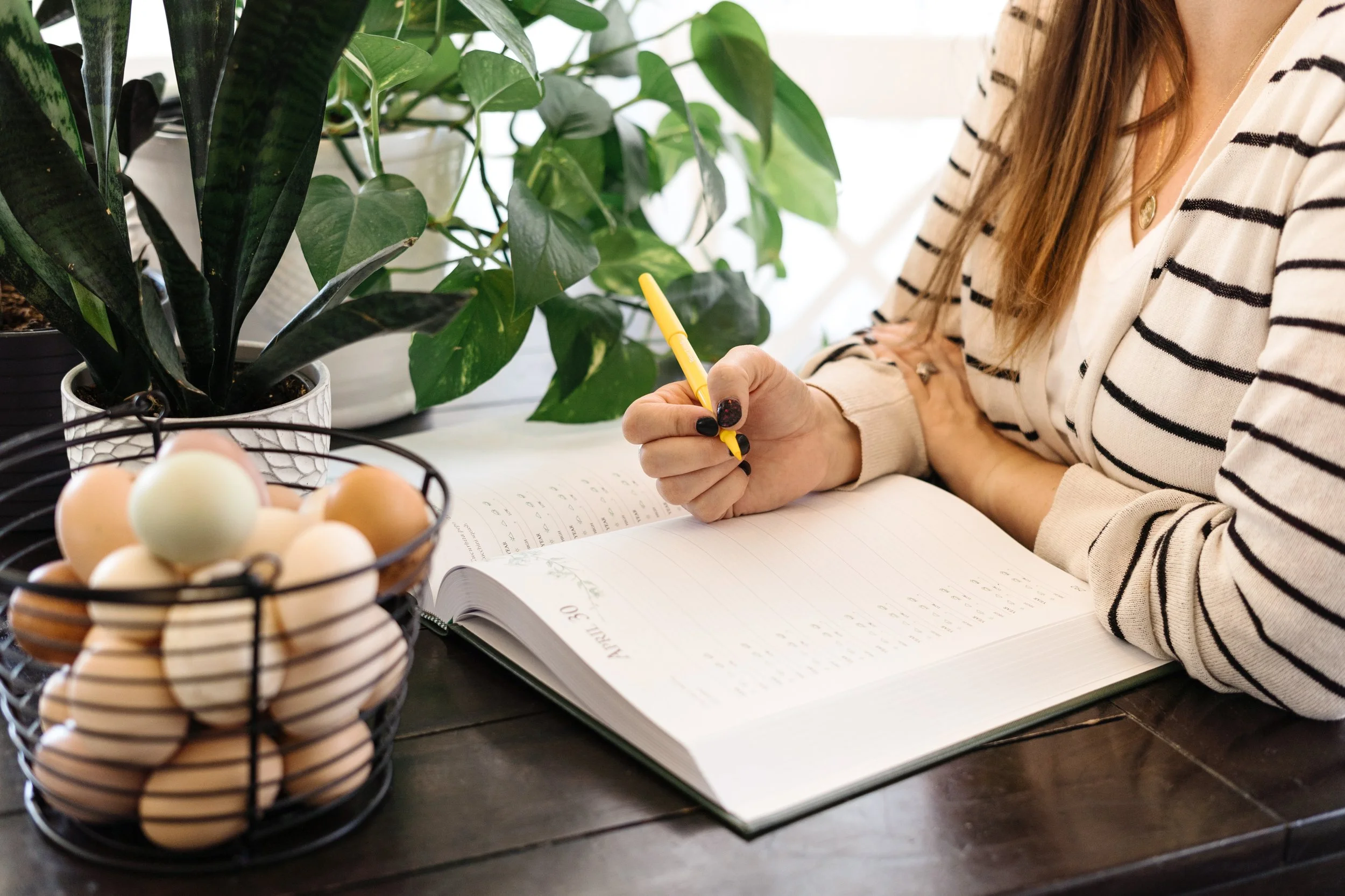 Woman writing in a dated paper planner with a yellow pen, surrounded by leafy green houseplants and a basket of multicoloured eggs on a table.