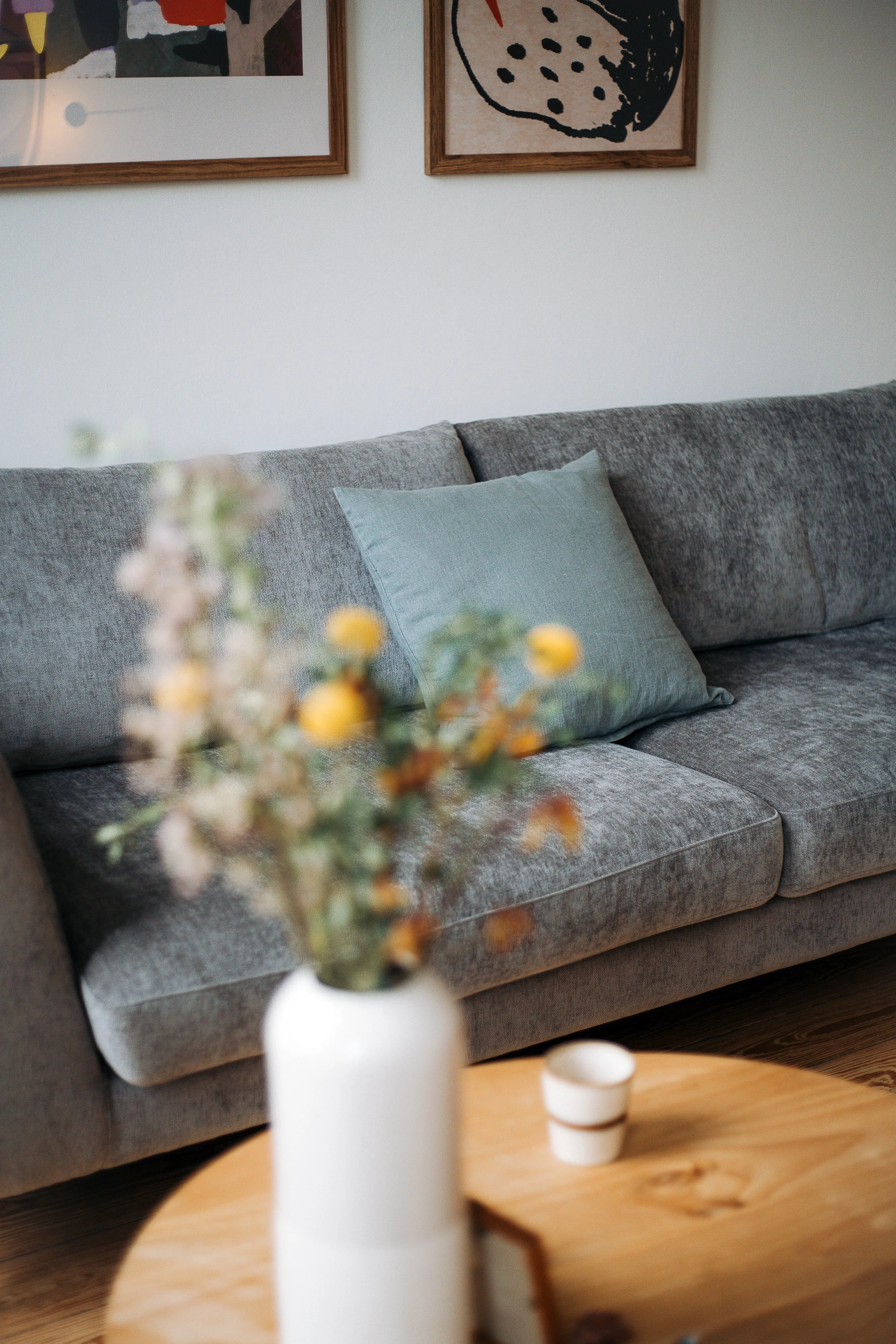 Gemütliches Wohnzimmer mit grauem Sofa, hellblauem Kissen, abstrakten Bildern an der Wand, Holz-Tisch mit Vase und Blumentopf