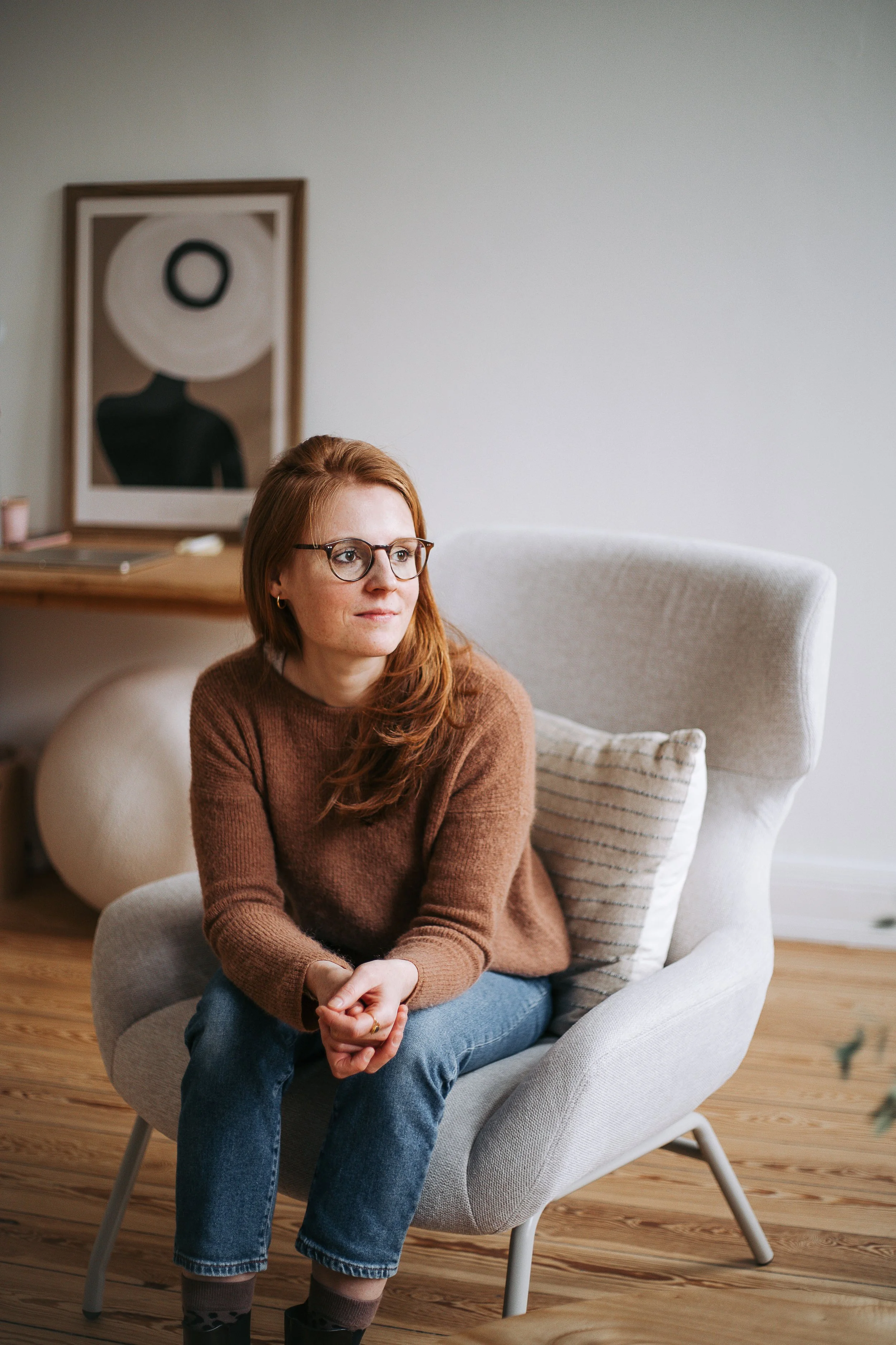 Eine Frau mit roten Haaren und Brille sitzt in einem weißen Ohrensessel, trägt einen braunen Pullover und blaue Jeans, im Hintergrund hängt ein abstraktes Artwork an der Wand.
