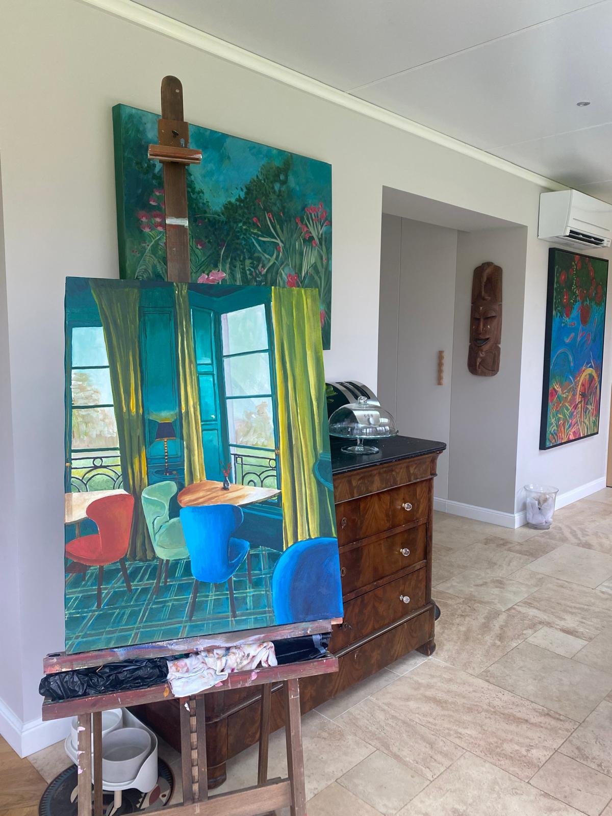 Chevalet avec une peinture colorée représentant un intérieur de salon, dans une pièce avec des œuvres d'art accrochées aux murs, une commode en bois, et un sol en pierre beige.
