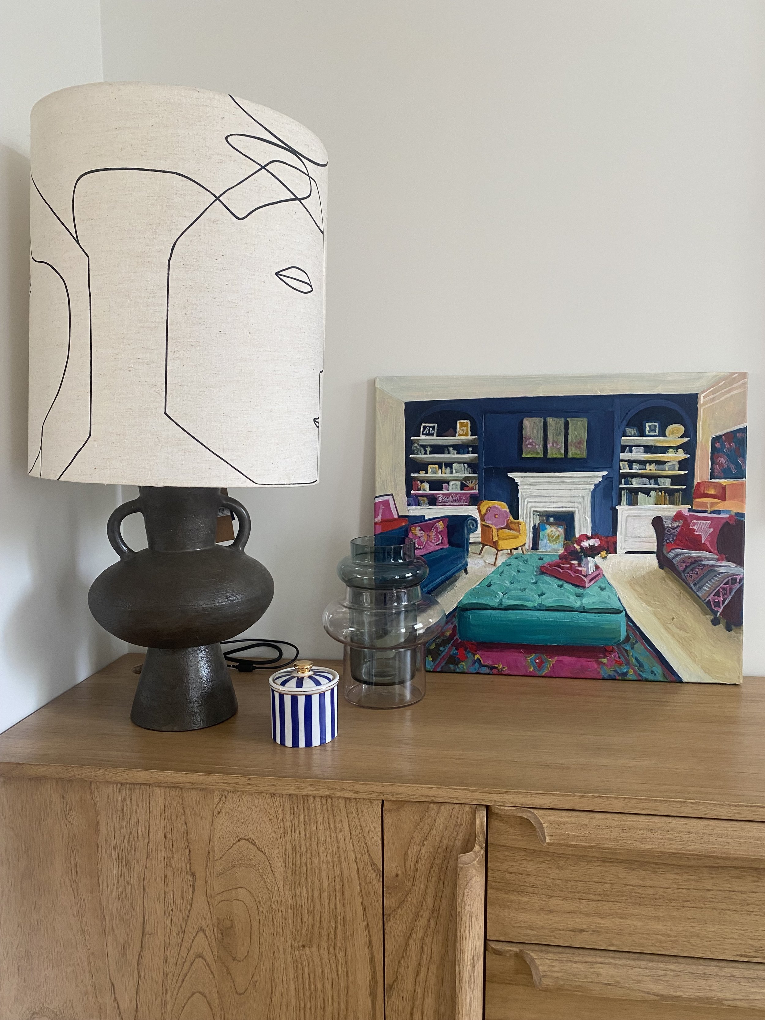Lampe décorative sur une commode en bois avec un vase en verre, une boîte rayée bleue et blanche et une toile colorée représentant un salon.
