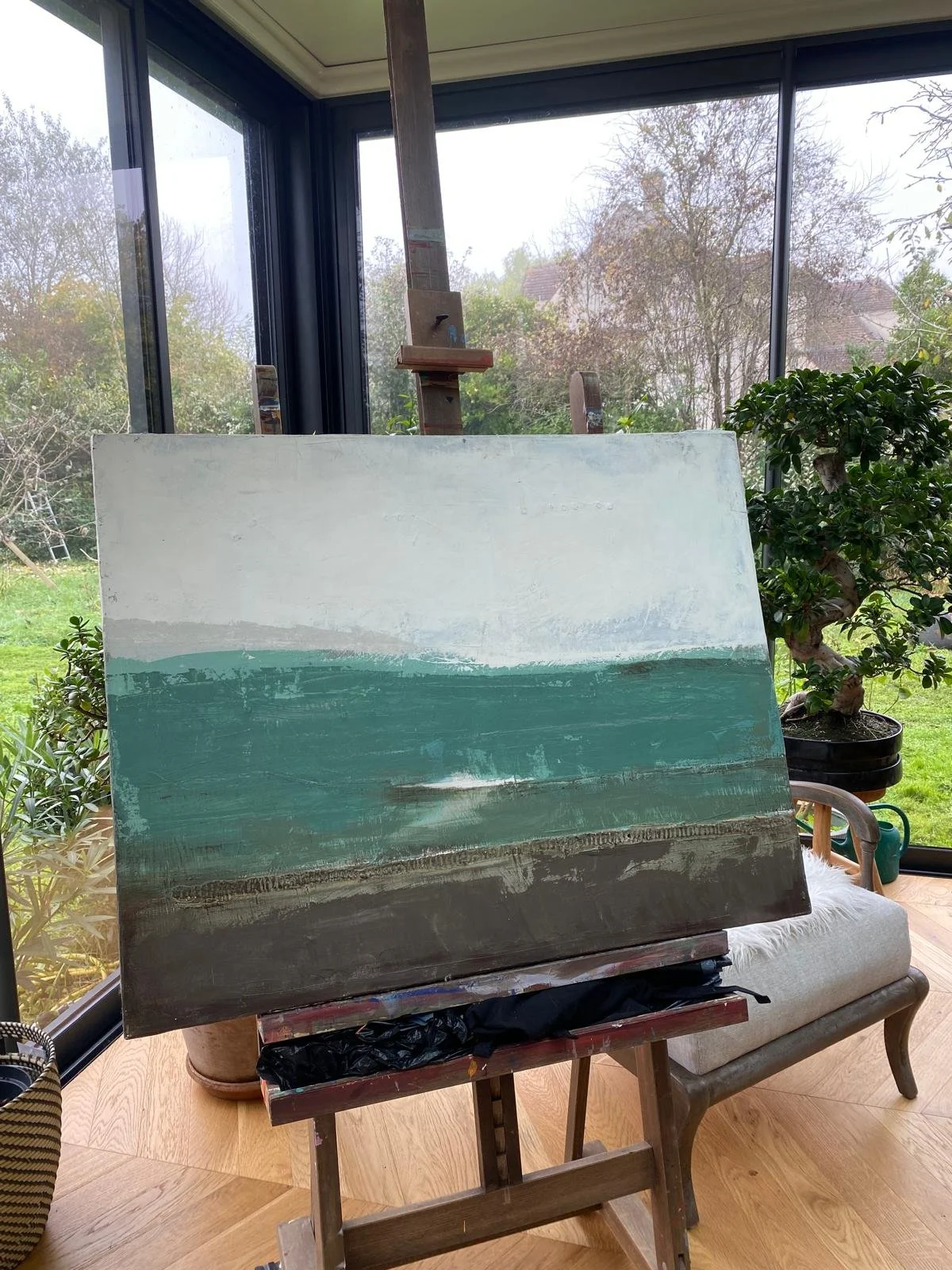 Tableau sur un chevalet dans une pièce avec baie vitrée, vue sur jardin verdoyant.