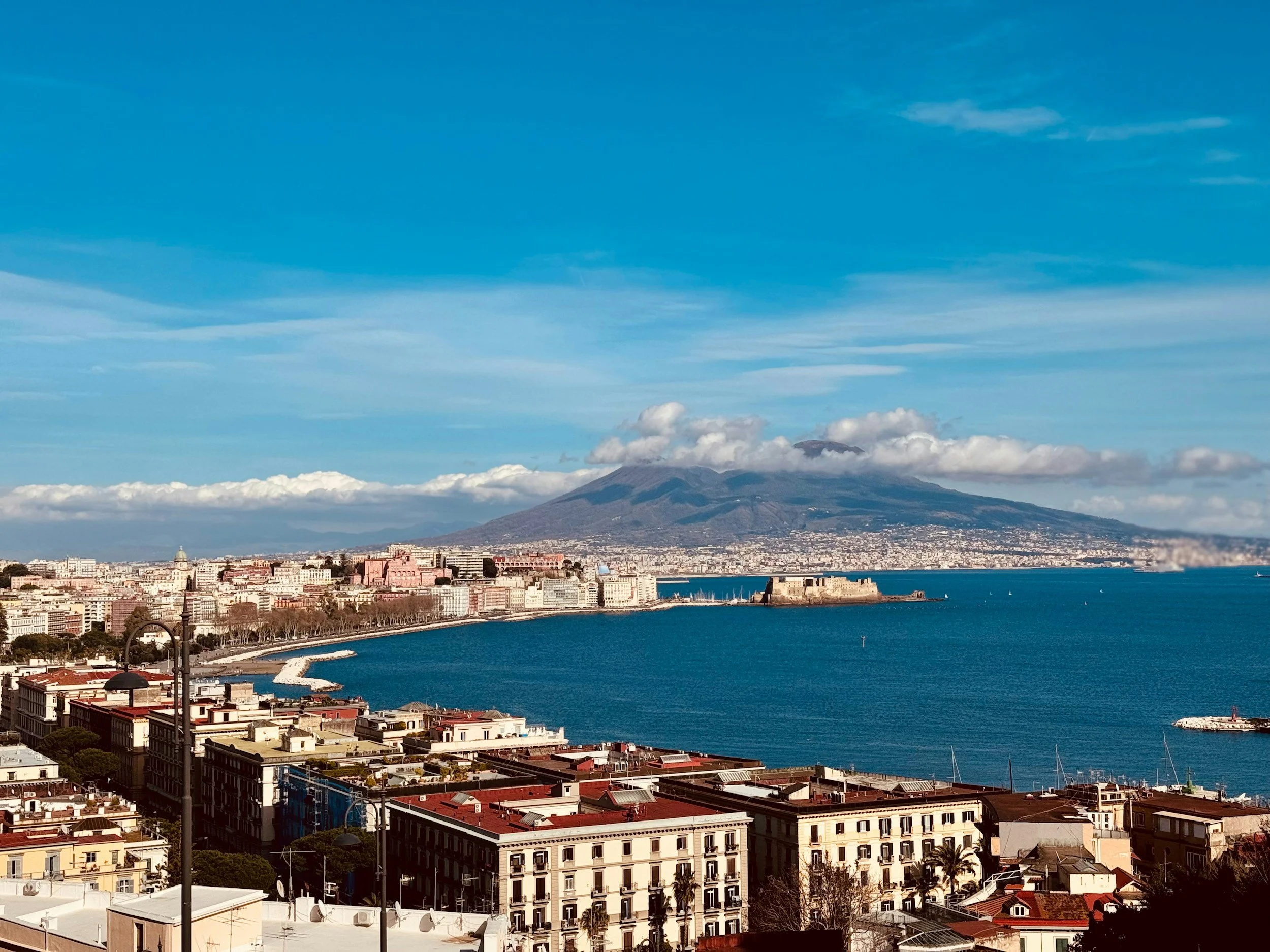 A cityscape view of Naples, Italy, with the Bay of Naples and Mount Vesuvius in the background on a clear day.