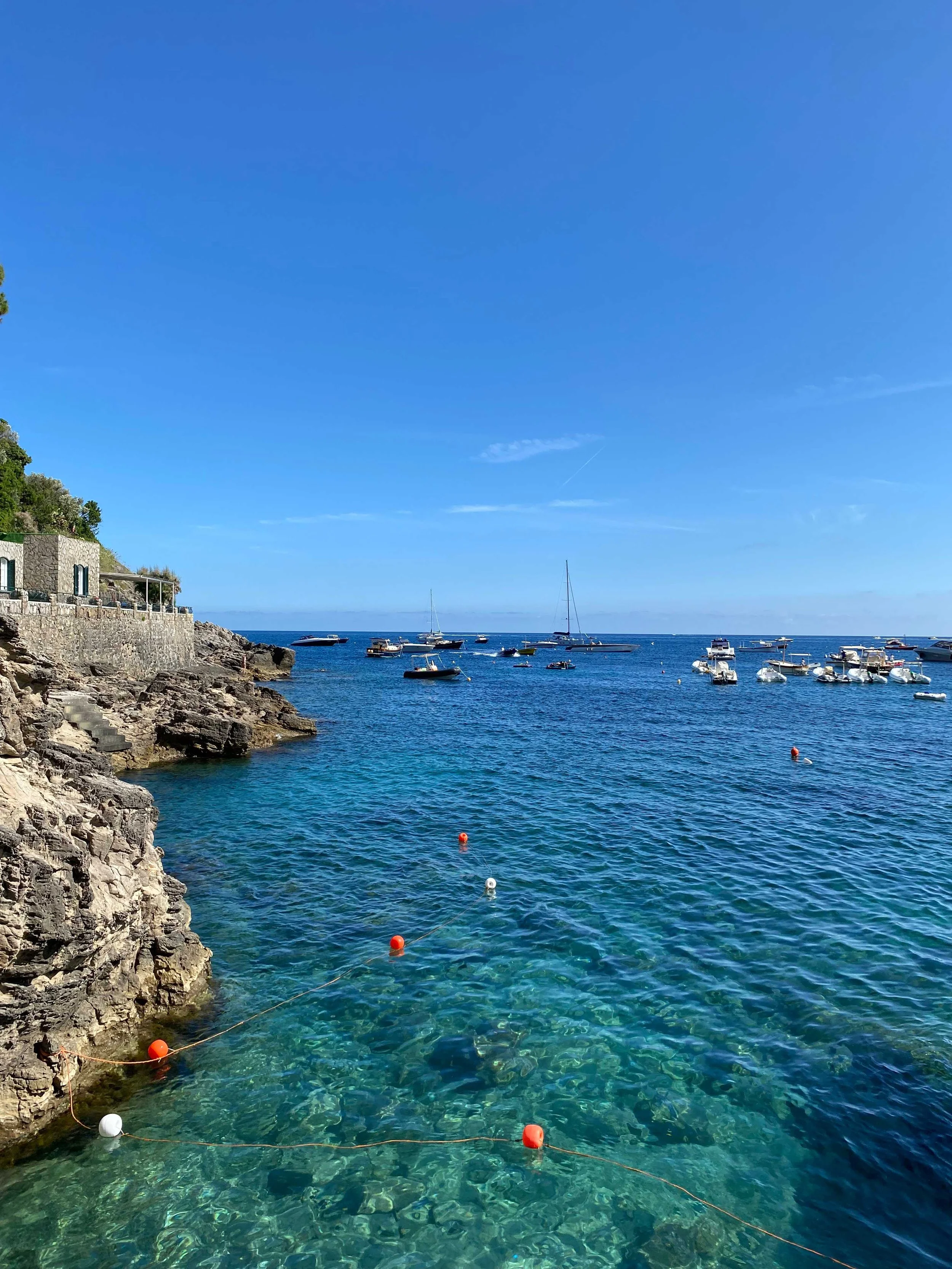 Coastal view with clear blue water, rocky shoreline, and small boats in the distance under a bright blue sky.