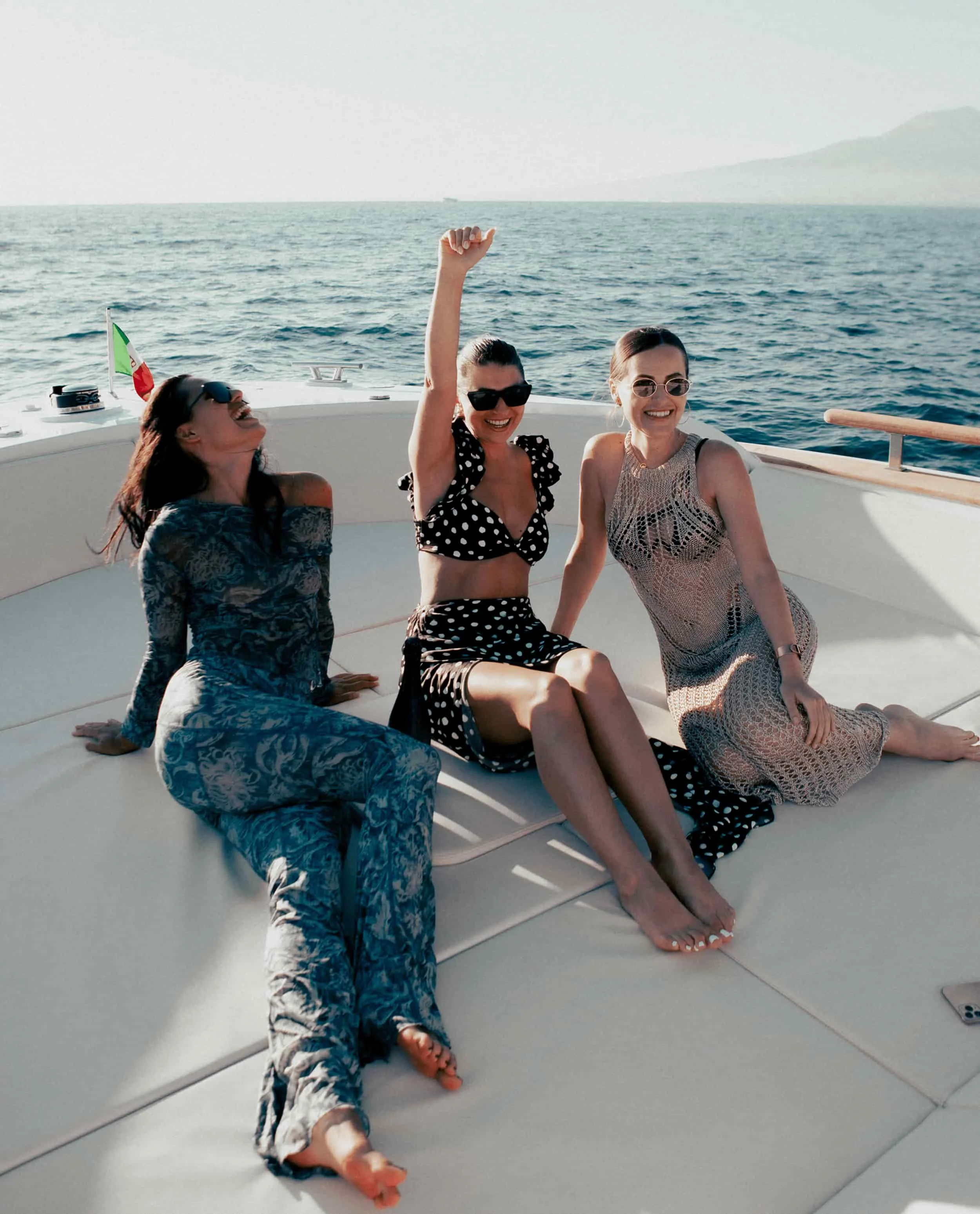Three women sitting on a boat deck, having fun, with ocean and distant mountains in the background.