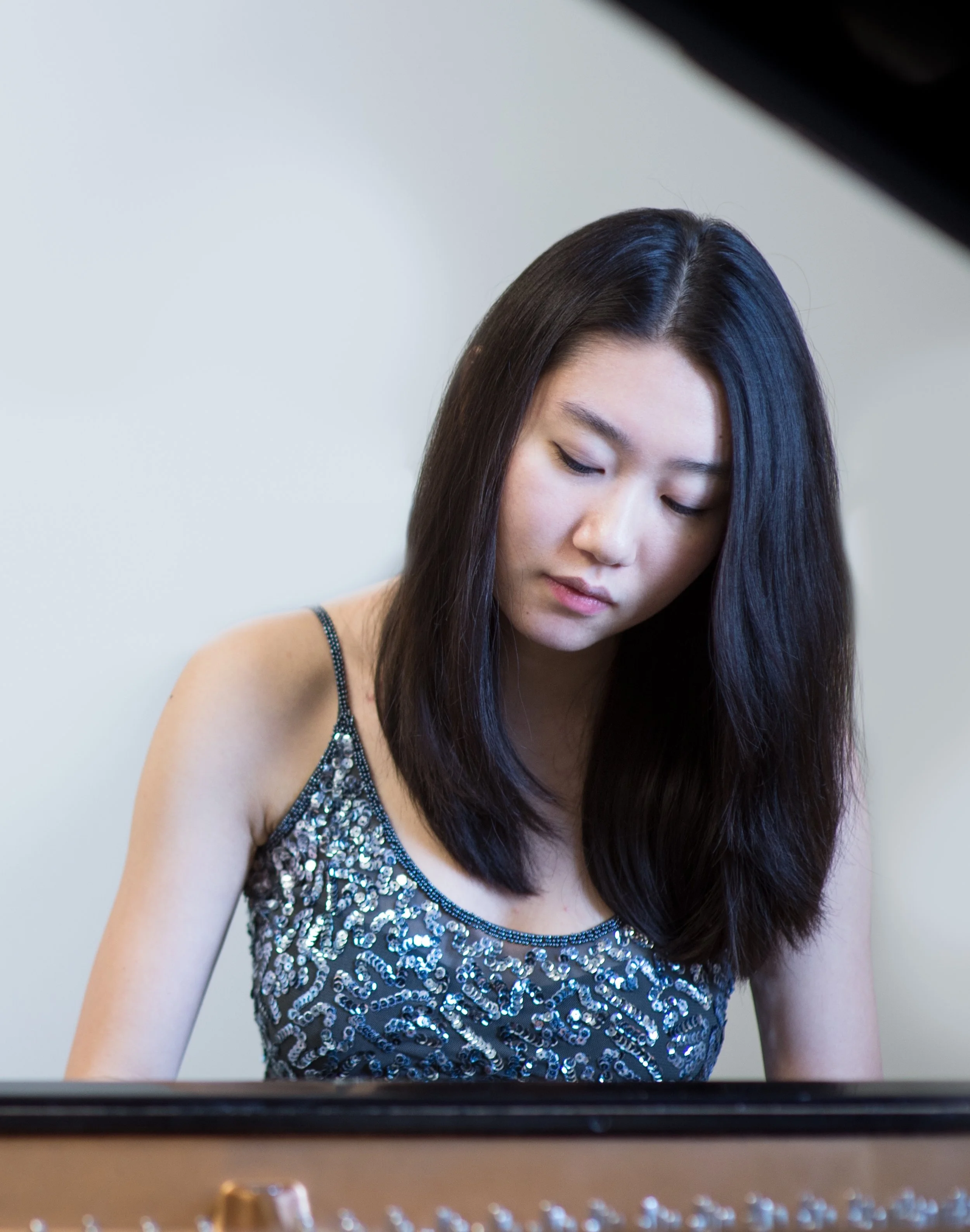 A young woman with long dark hair and fair skin is looking down, wearing a sleeveless dress decorated with shiny sequins, in front of a plain light-colored wall.