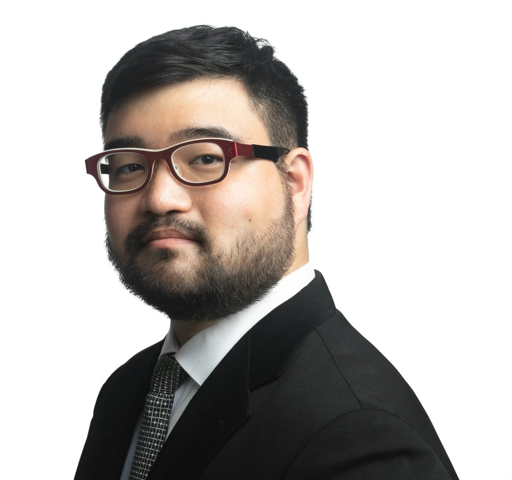 Partial portrait of a man with black hair, glasses, and a beard, dressed in a tuxedo with a checkered tie, against a white background.