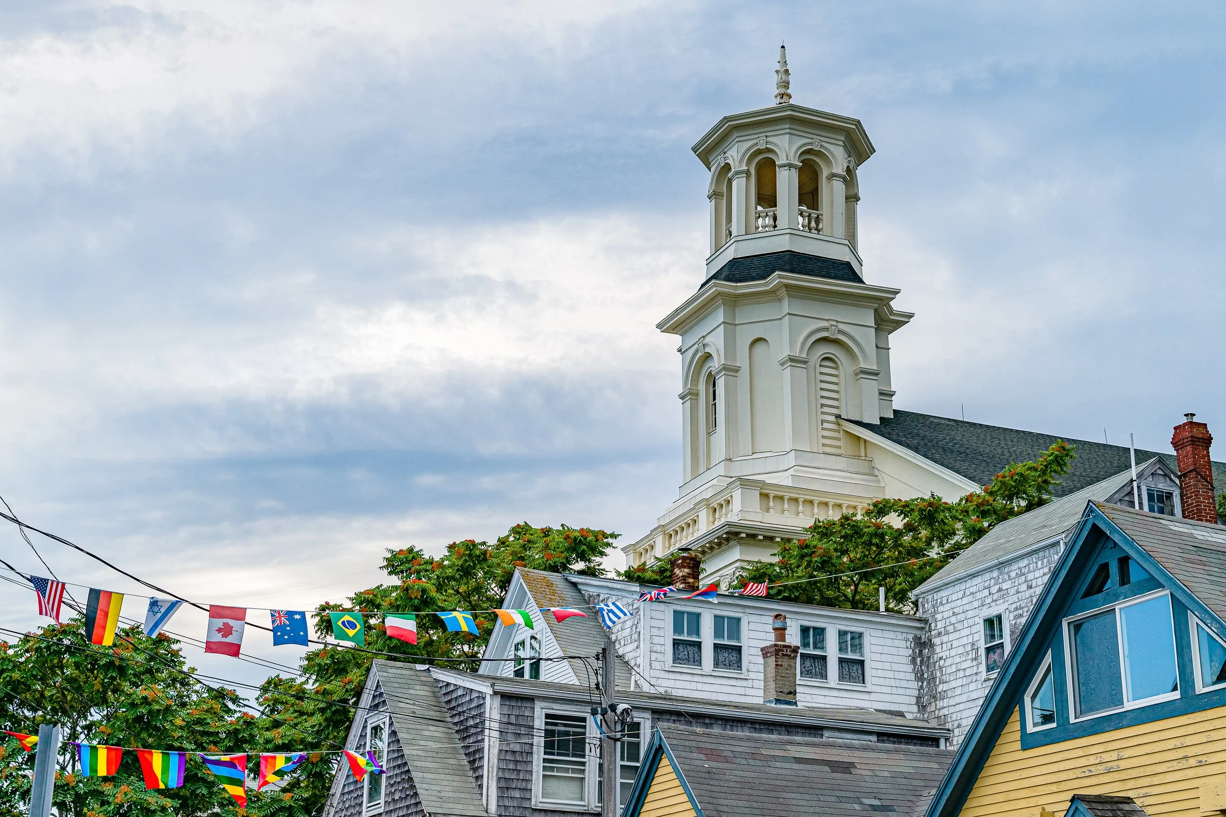 A church tower with a white facade and a cupola on top, surrounded by houses with colorful rooftops and international flags hanging above.