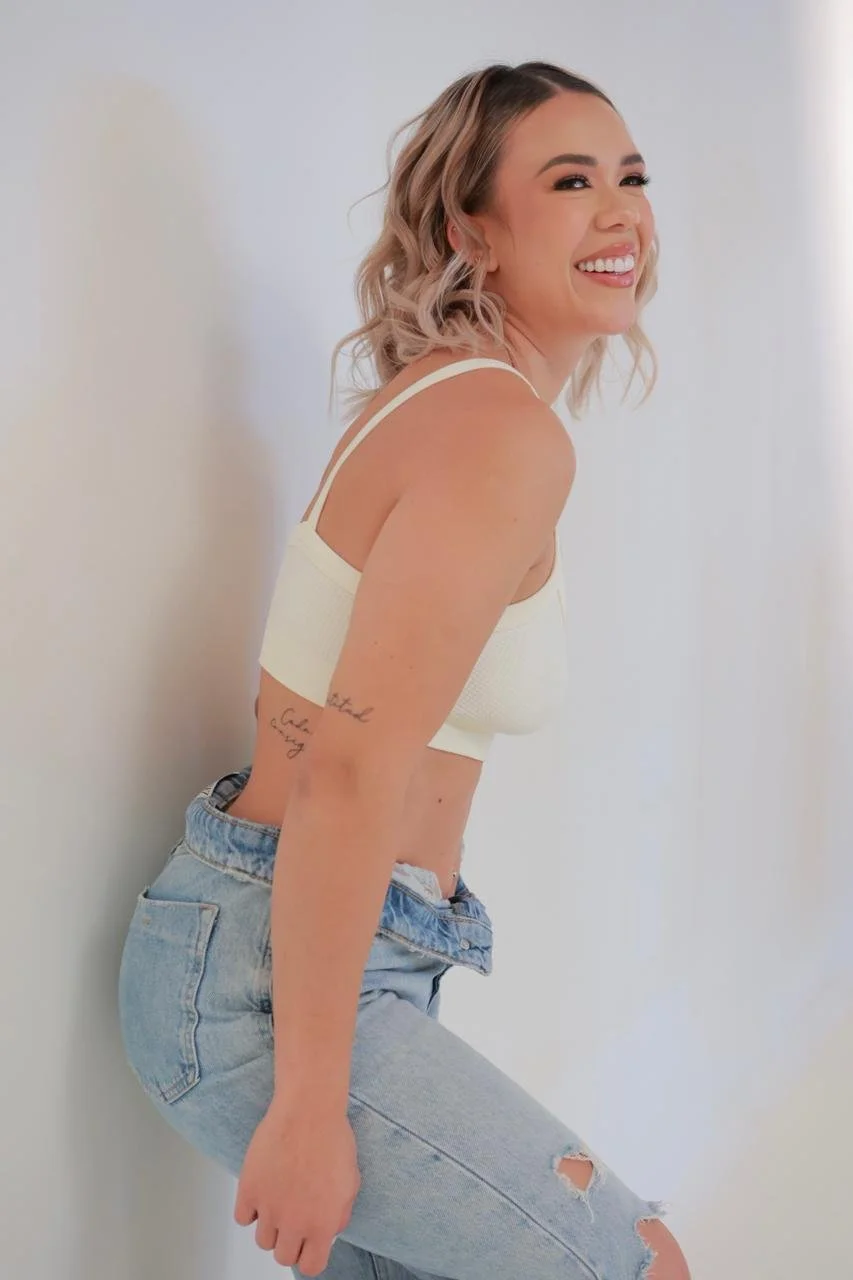 Joven mujer sonriendo, con cabello rubio y rizado, usando camiseta blanca y jeans rotos, posando inclinada a la pared blanca.