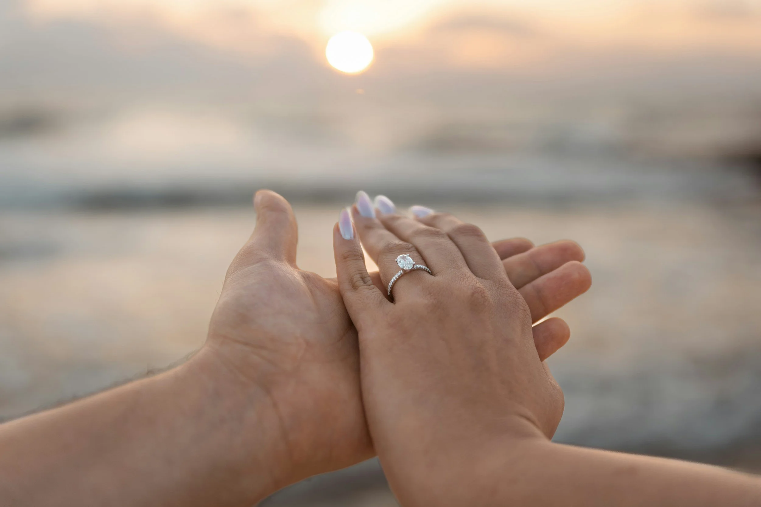 Close-up of two hands holding each other at the beach during sunset, with a wedding ring visible on the ring finger of the hand on top.