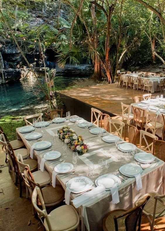 A beautifully set outdoor dining table decorated with a floral centerpiece, surrounded by chairs, situated near a natural water feature, under a canopy of trees.