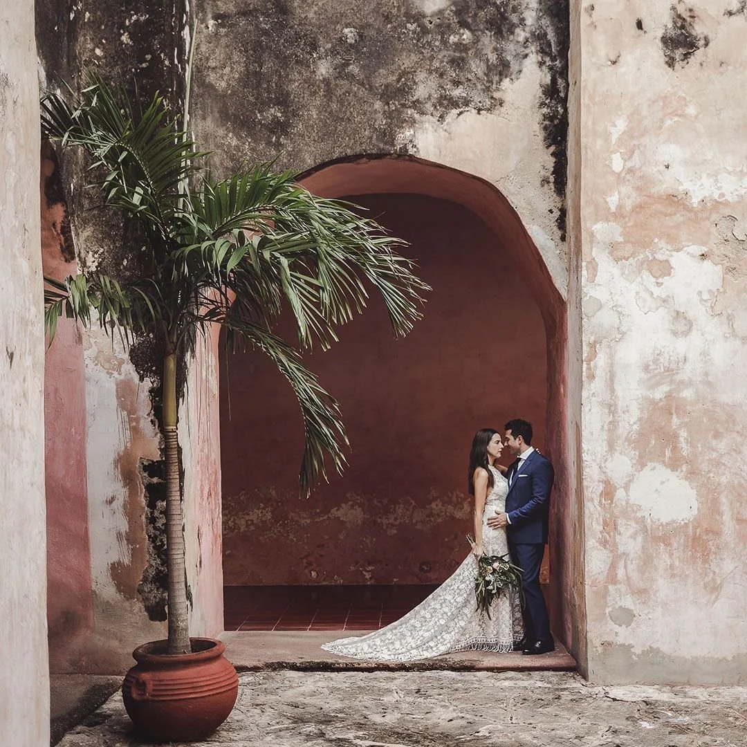 A bride and groom standing close together, facing each other, under an archway with textured pink walls, a potted palm tree on the left, and a rustic, weathered background.