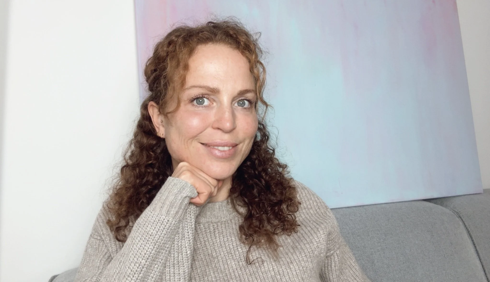 Eine junge Frau mit lockigem braunem Haar, blauen Augen und einem freundlichen Lächeln, sitzt auf einem grauen Sofa vor einer weißen Wand mit einem großen, mehrfarbigen Kunstwerk im Hintergrund.