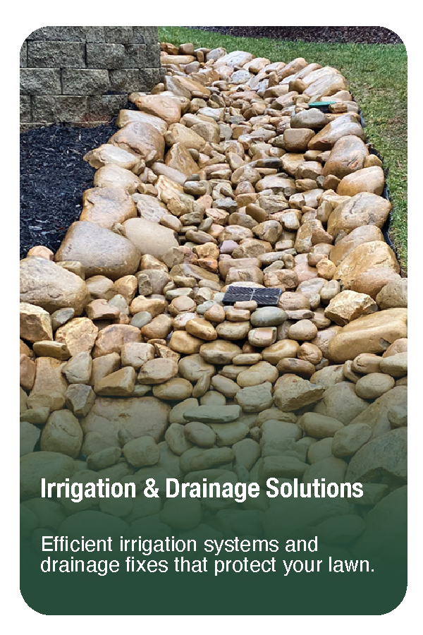 A garden landscape with a stone-lined drainage bed filled with a variety of smooth, rounded rocks, surrounded by green lawn and brick wall.