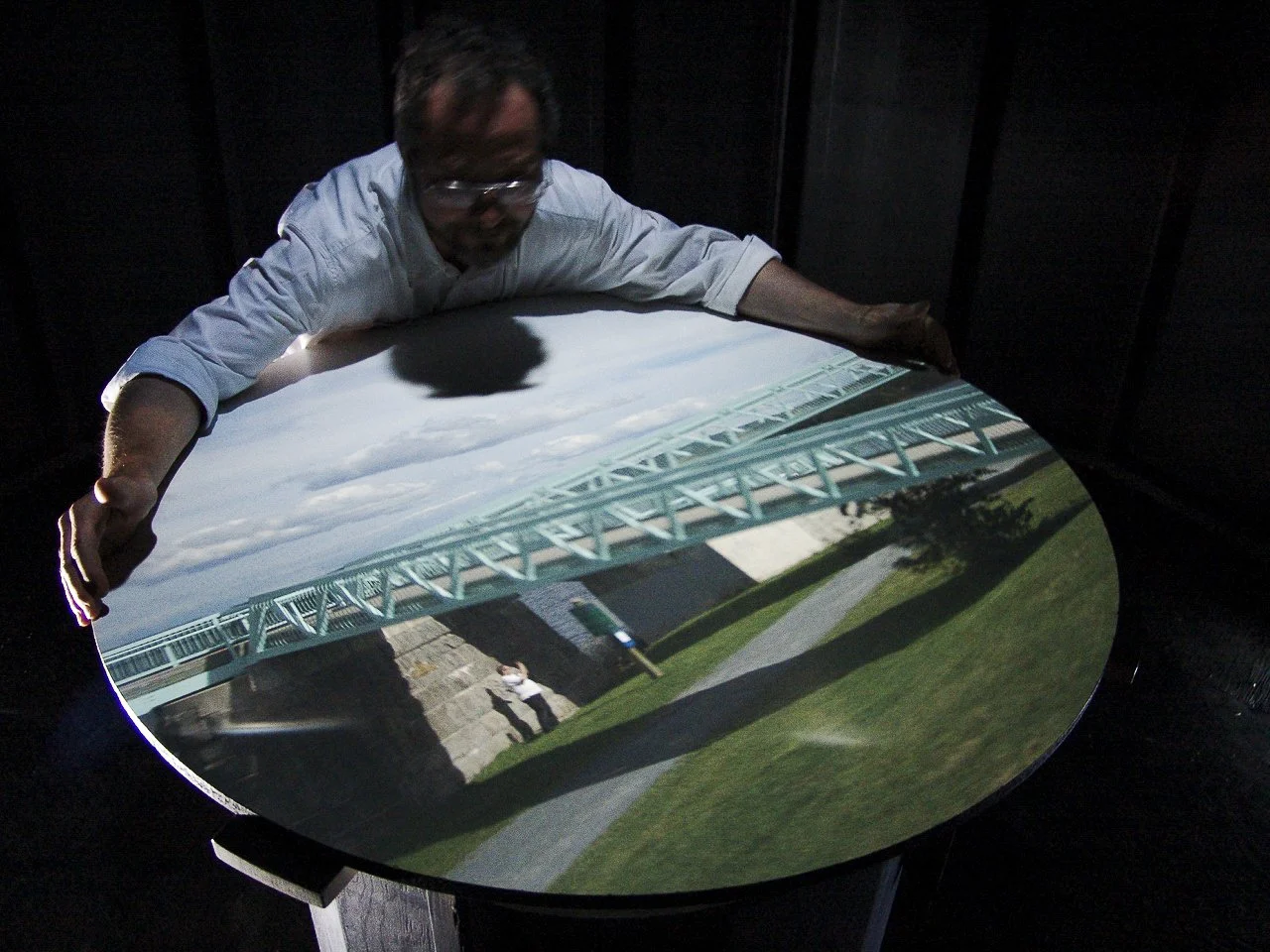 Man adjusting camera obscura projection of a bridge and landscape on a round table, with dim lighting.