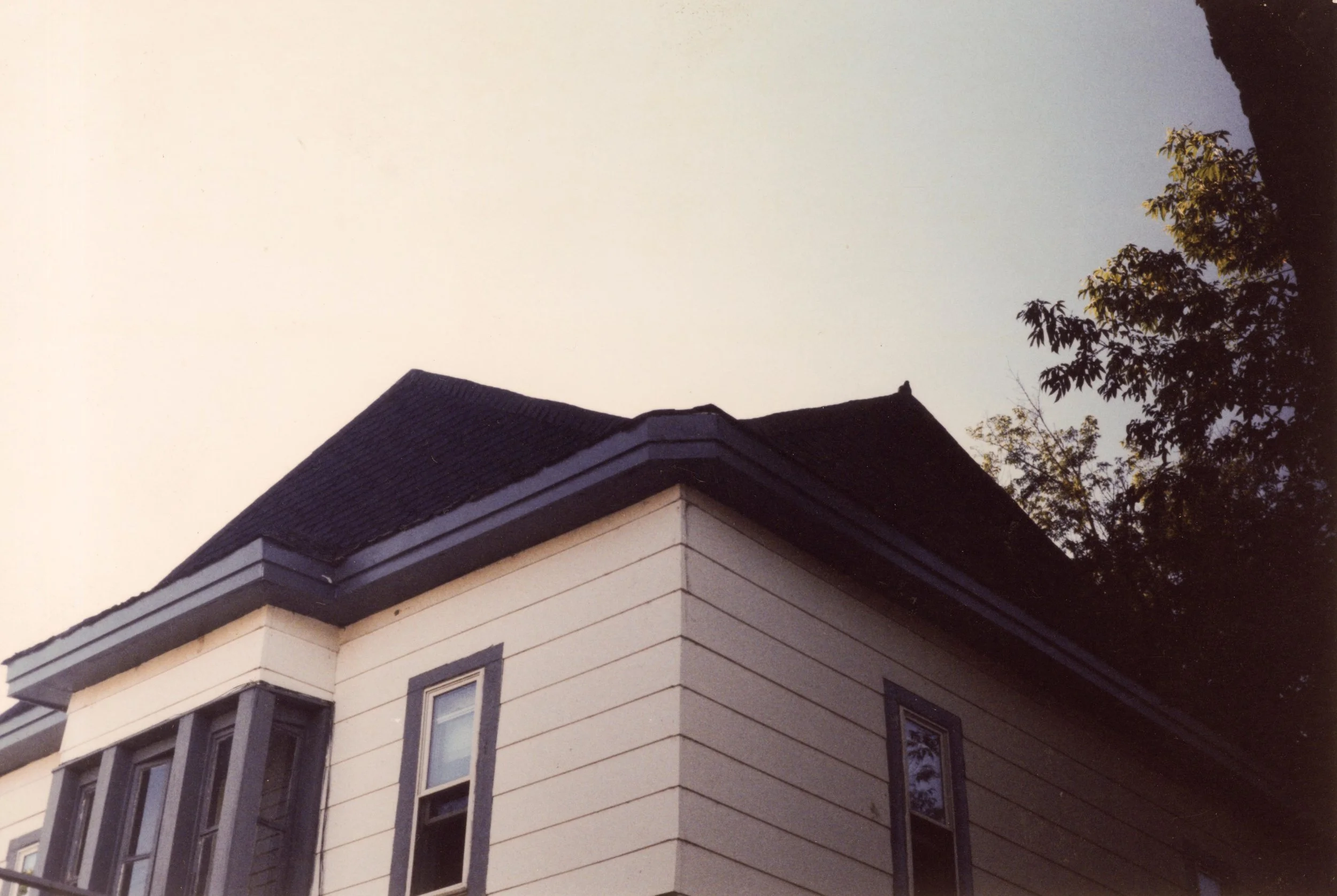 Corner view of a vintage house with a dark roof, beige siding, and white-framed windows, surrounded by trees.