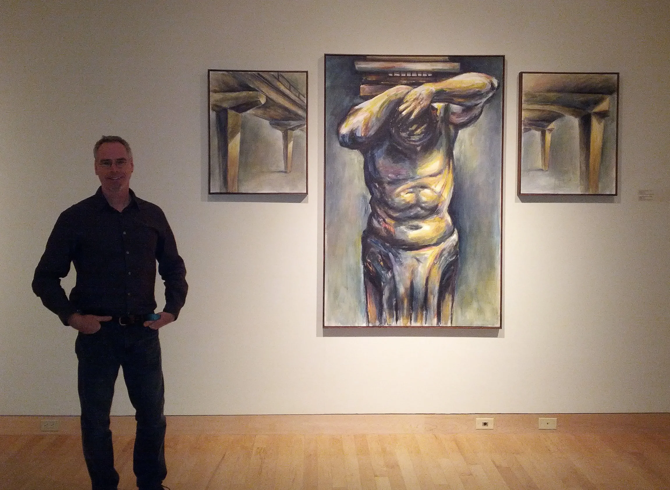 Man standing beside a set of three abstract paintings displayed on a gallery wall.
