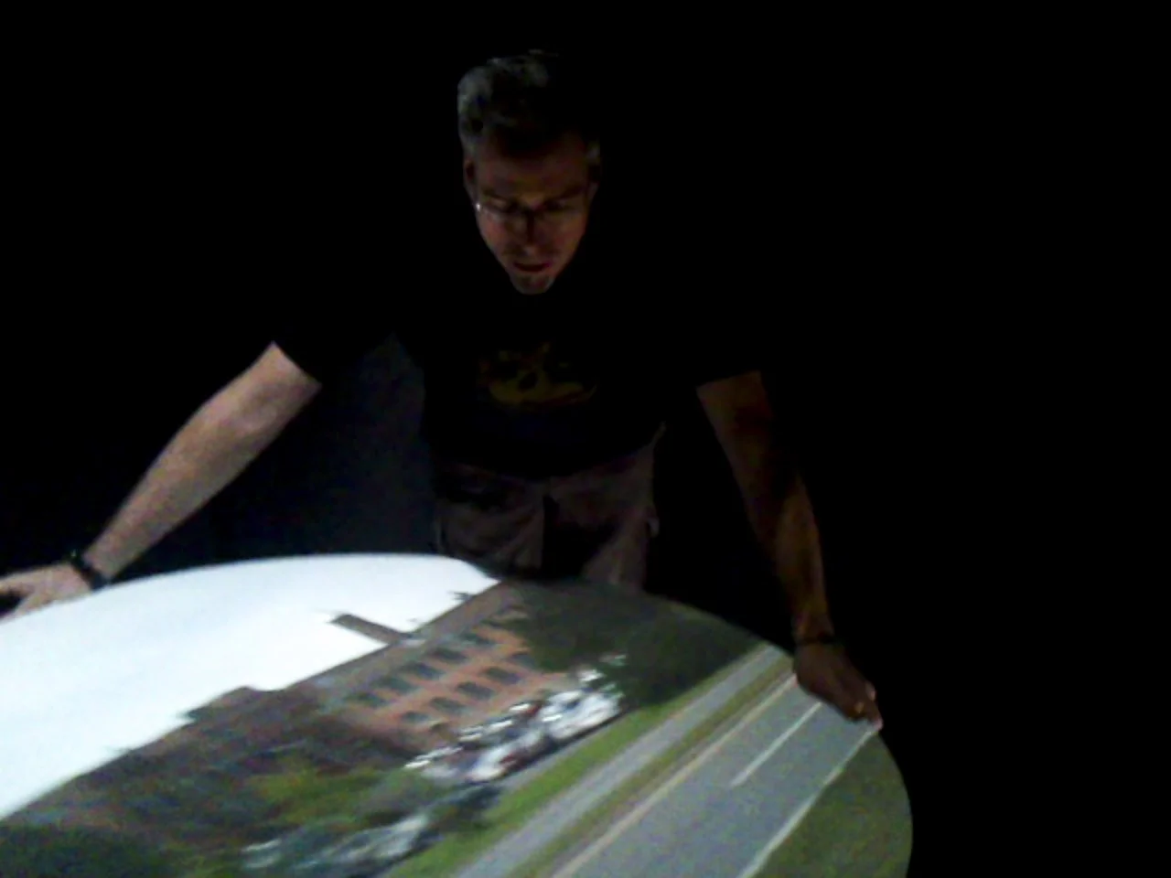 A person standing over a circular table displaying a projected aerial image of a building and road, in a dark room.