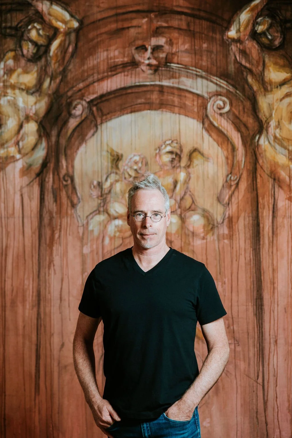 Man in black T-shirt standing in front of an artistic mural with cherubs.