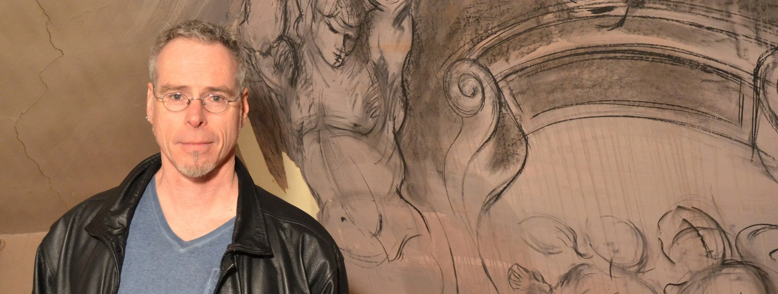 Man wearing glasses and a black leather jacket standing in front of a large, detailed drawing on a wall featuring figures and intricate designs.