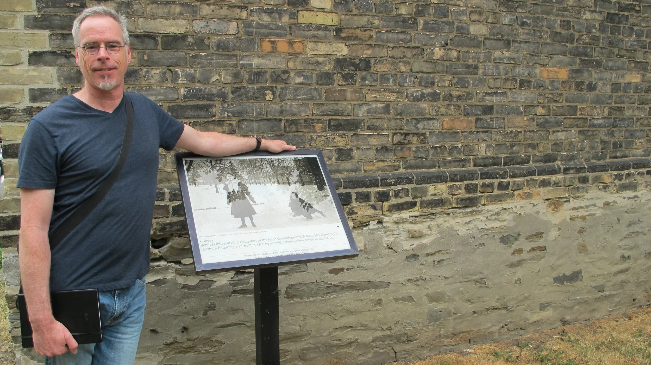 A man with gray hair and glasses stands beside an informational plaque mounted on a post against a brick wall. He is wearing a blue T-shirt and holding a black tablet.