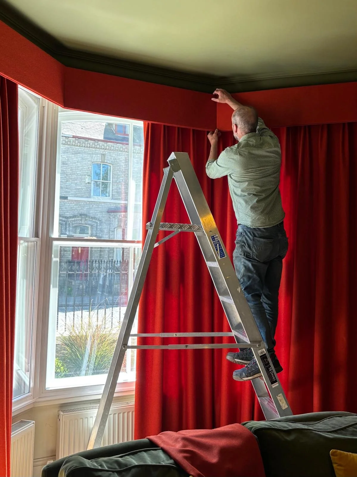 A curtain fitter in York installs a pelmet above a high bay window. The fabric is a bold red.