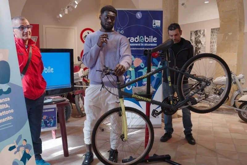 Tre uomini presentano una bicicletta all'interno di un'installazione o mostra, con uno dei uomini che tiene la bicicletta sollevata da terra. La scena include uno sfondo con pannelli informativi e display. Uno dei uomini indossa una felpa grigia e pa