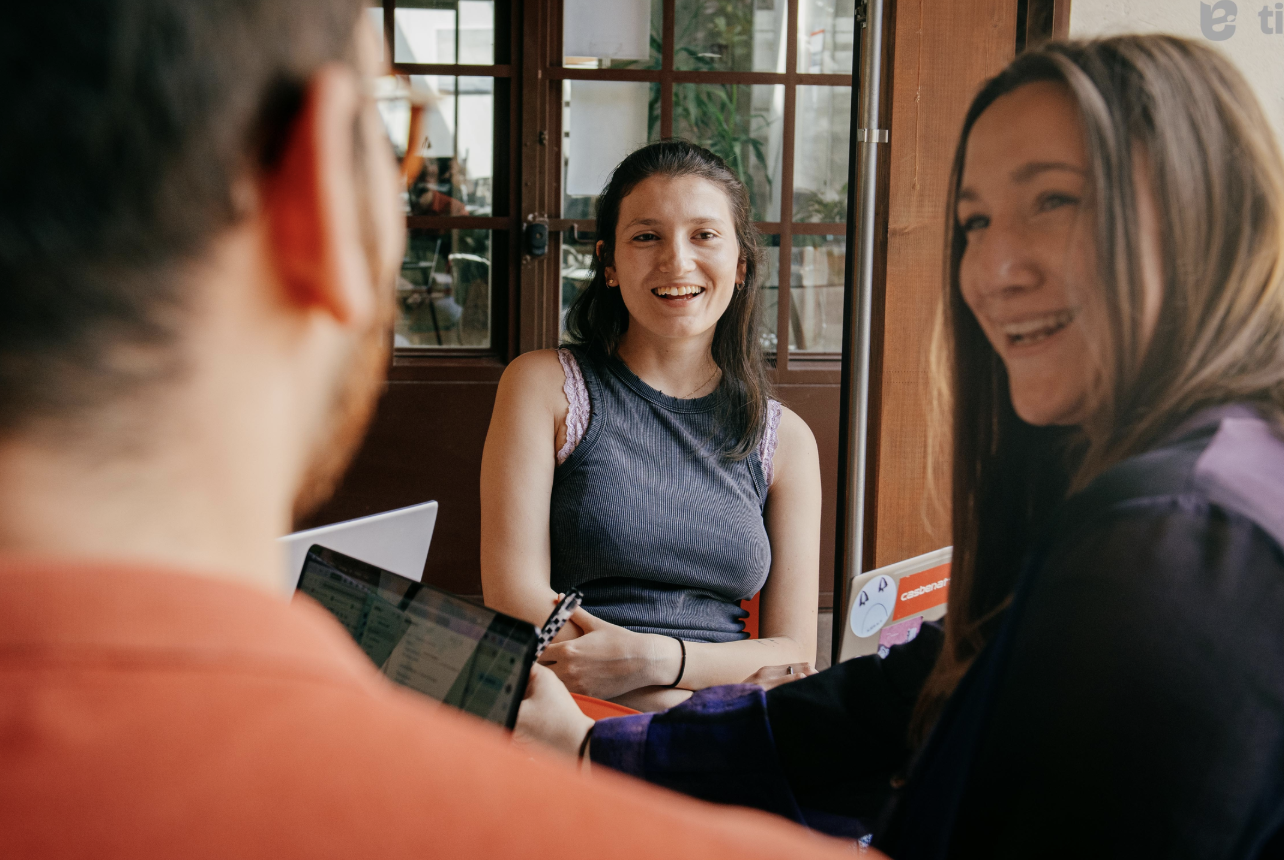 Tre persone sedute a un tavolo che conversano e sorridono, in un ambiente luminoso con vetrate alle spalle.