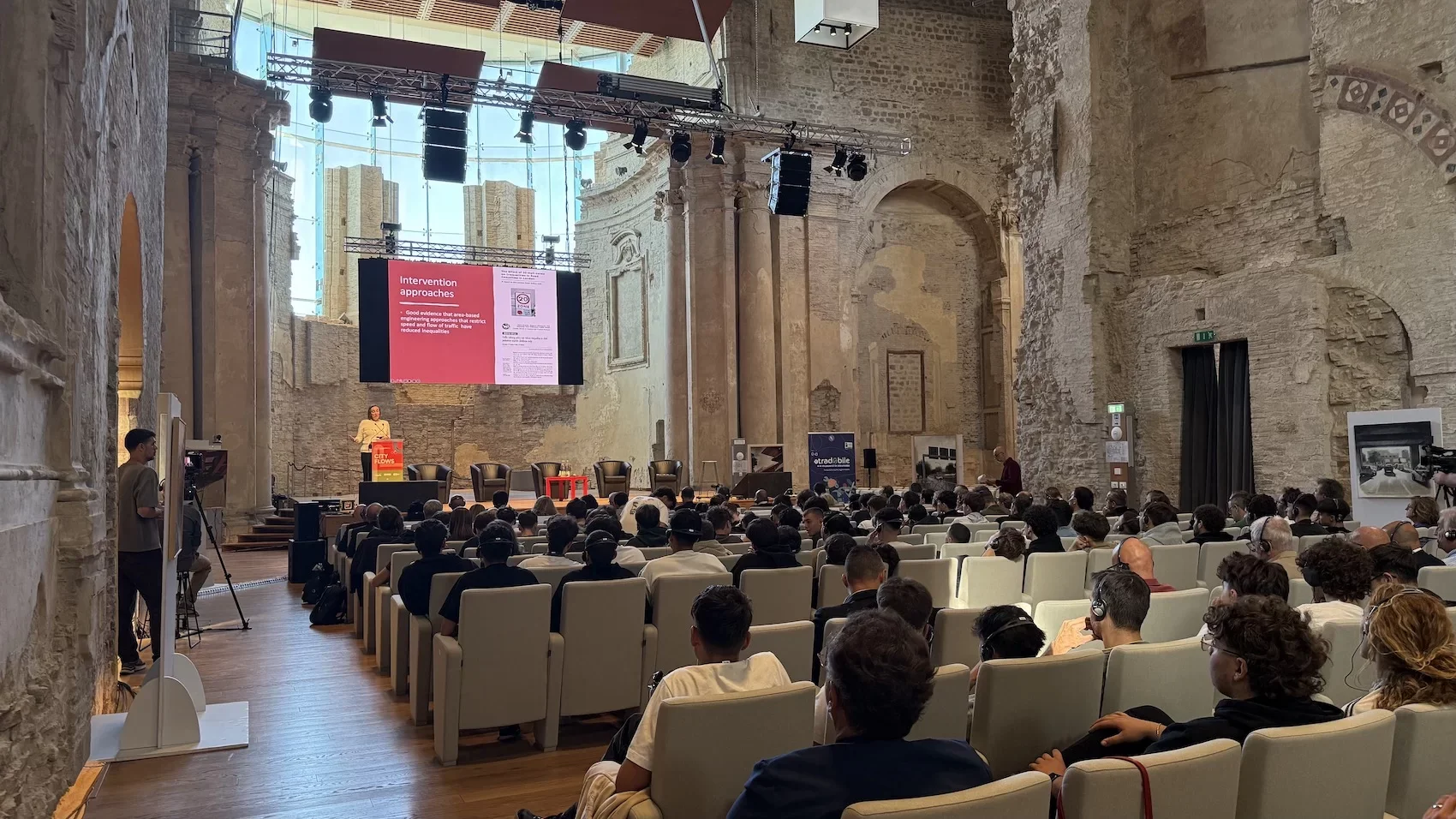 Una conferenza si svolge in un grande spazio con pareti di pietra antiche e un soffitto di vetro. Sul palco ci sono diverse sedie e un podio, con una donna che tiene una presentazione su uno schermo grande. L'audience, composta in gran parte da giova