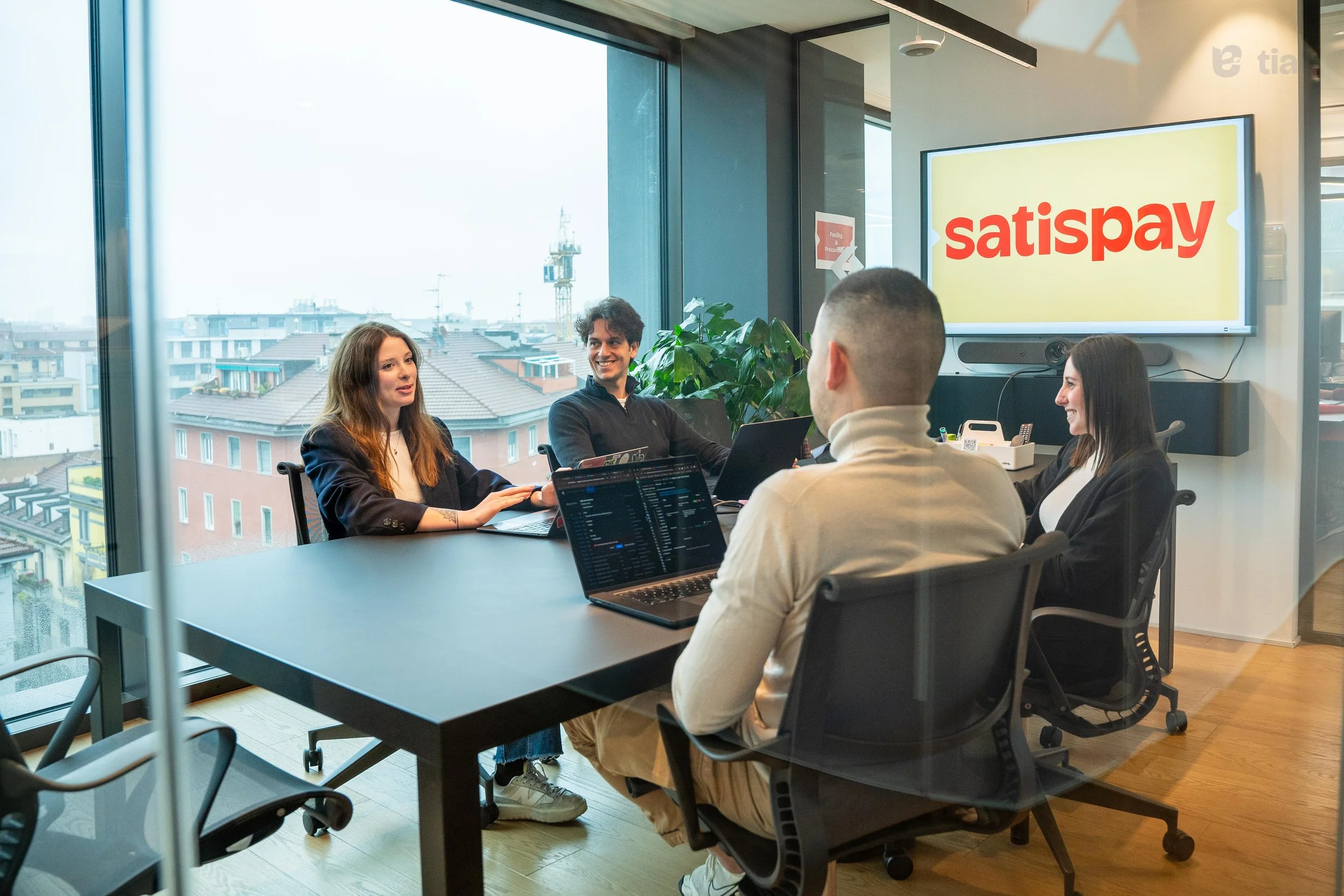 Cinque persone sedute intorno a un tavolo in una sala riunioni con grandi finestre che mostrano un panorama urbano. Sullo sfondo, uno schermo con la scritta "satispay" in rosso su sfondo giallo.