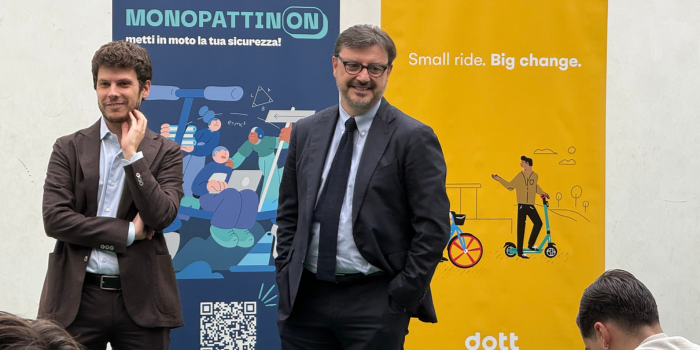 Due uomini sorridenti in piedi davanti a due poster pubblicitari, uno blu e uno giallo, che promuovono l'uso di monopattini e biciclette per la sicurezza e il trasporto sostenibile.