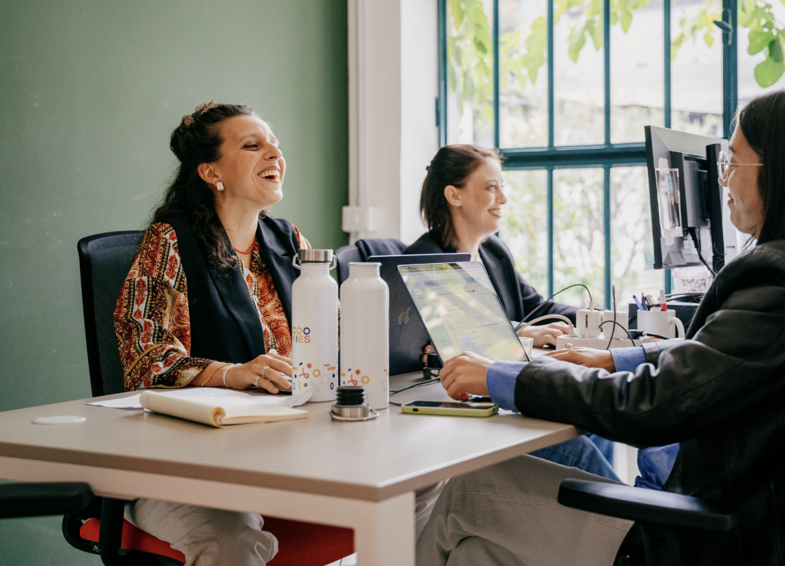 Tre persone sorridenti sedute a una scrivania in un ufficio, due di loro davanti a computer e una che sorride con una bottiglia d'acqua sulla scrivania.