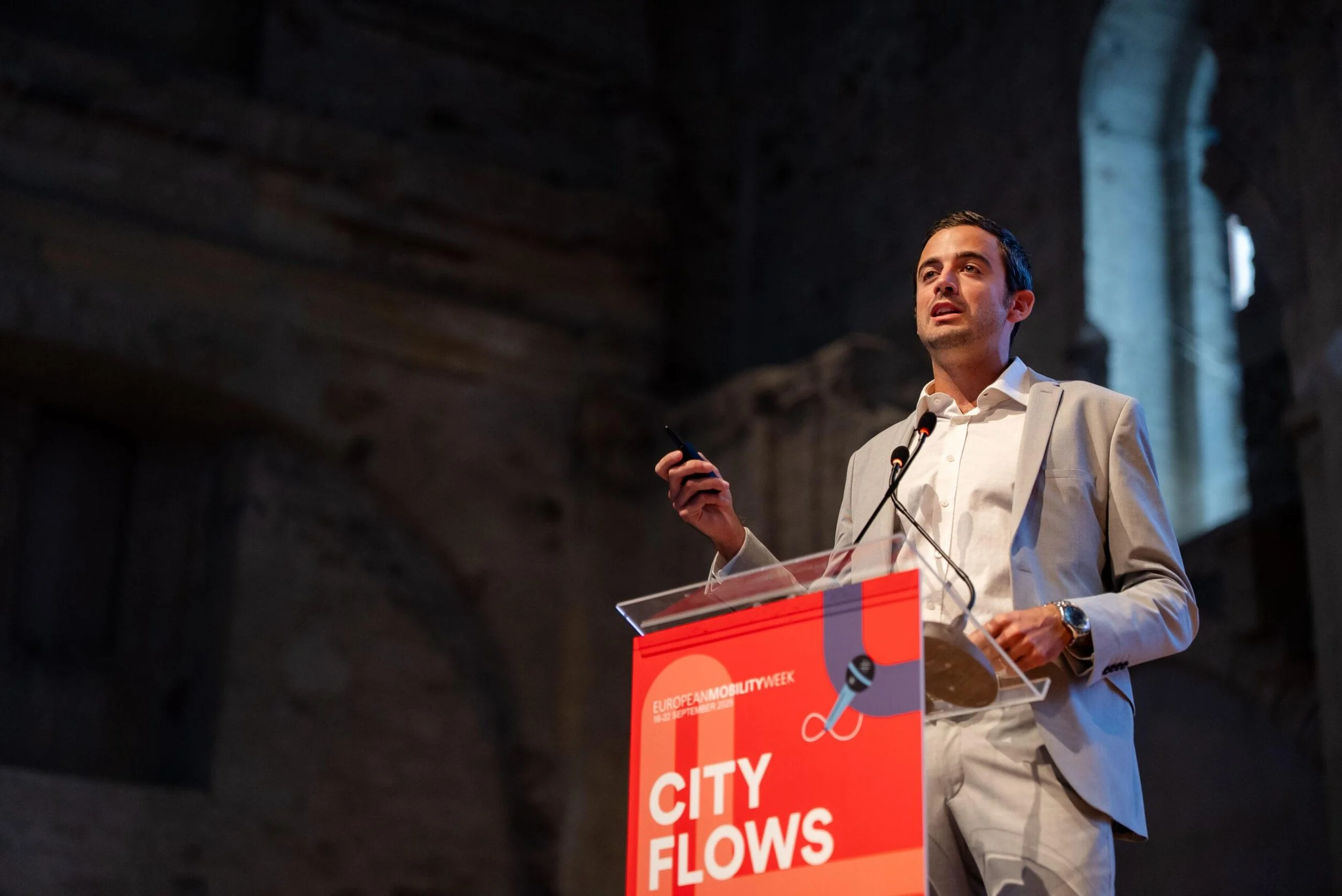 Uomo che tiene un microfono e un dispositivo mobile durante una conferenza. In piedi dietro un podio con il logo 'City Flows'. Sfondo di un edificio storico con archi e finestre.